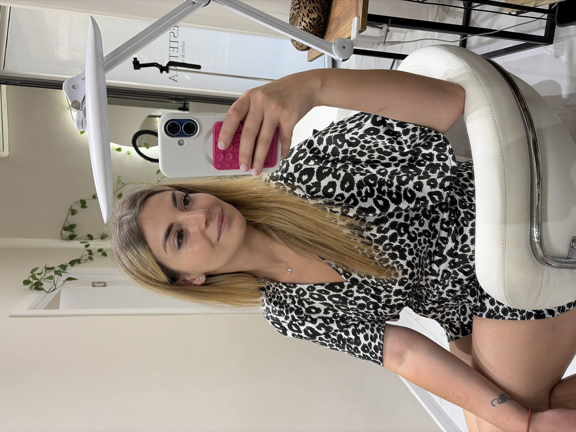 A woman with blonde hair is sitting in a white chair, taking a selfie in a room that appears to be a salon. She is wearing a black and white leopard print dress. A ring on her left hand is visible, and a delicate necklace with a clear stone rests on her chest. Her right arm is resting on the armrest of the chair, and her phone, with a pink popsocket, is held in her right hand. In the background, a white lamp with an adjustable arm is visible, along with some greenery hanging from the wall. A blurred reflection in a glass surface shows the word "ESTETIKA" and "salon" written backwards. The overall setting suggests a commercial establishment, likely a beauty salon, possibly located in Budva, Montenegro.