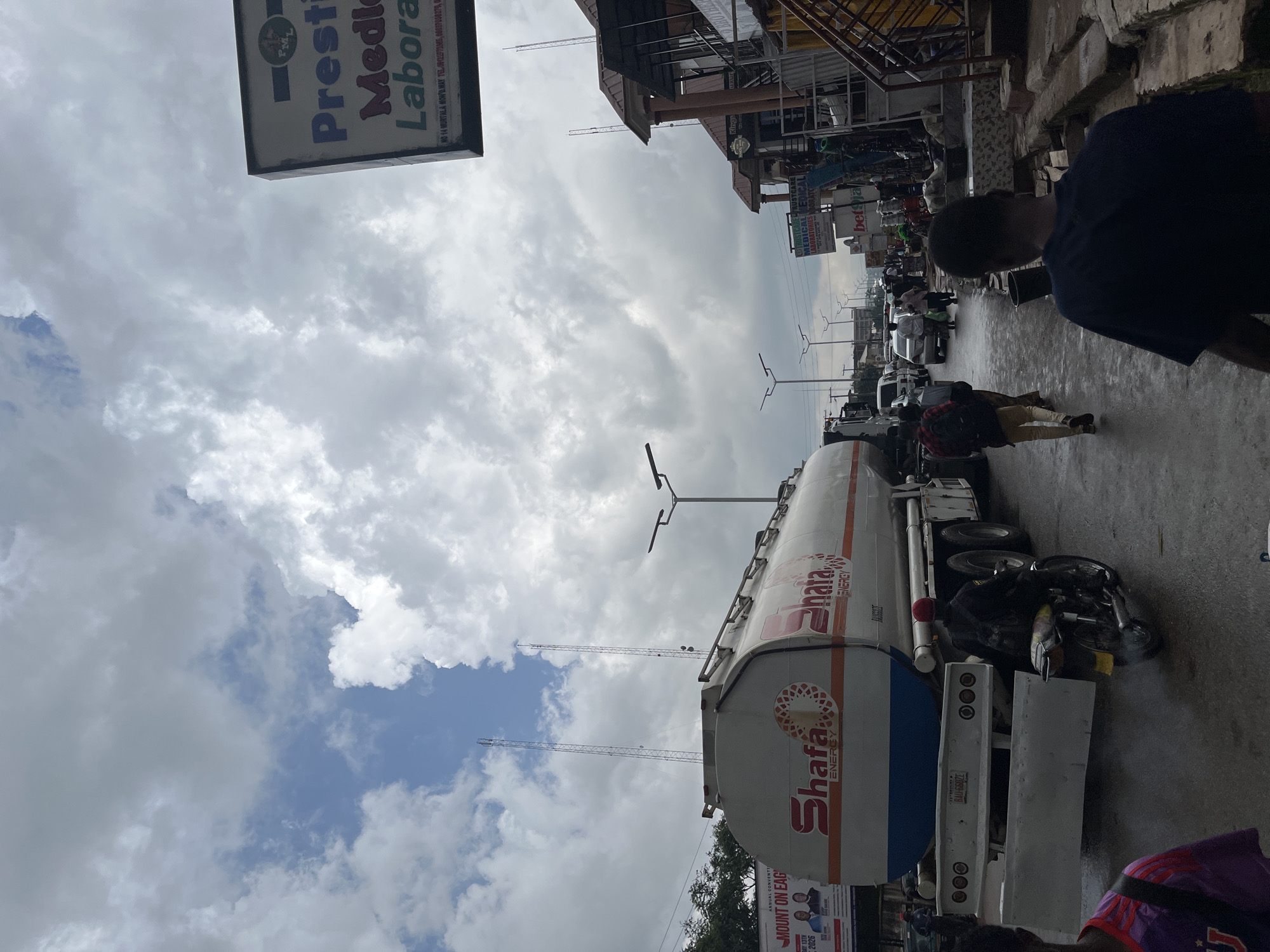 In Jos, Nigeria, a cloudy sky looms over a street scene. A large tanker truck, branded with "Shafa ENERGY," is parked on the wet asphalt, suggesting recent rain. The truck has a white body with orange and blue accents. To the right of the truck, a man wearing a red cap rides a motorcycle, exhaust fumes visible. Several other vehicles, including cars and a white van, are present on the road, indicating moderate traffic.
On the right side of the street, buildings line the road with awnings and signs, some appearing to be shops or businesses. One prominent sign reads "Prestige Medical Laboratories." People are walking along the street and sidewalk, some in groups, others singly. The overall atmosphere is that of a busy urban street.