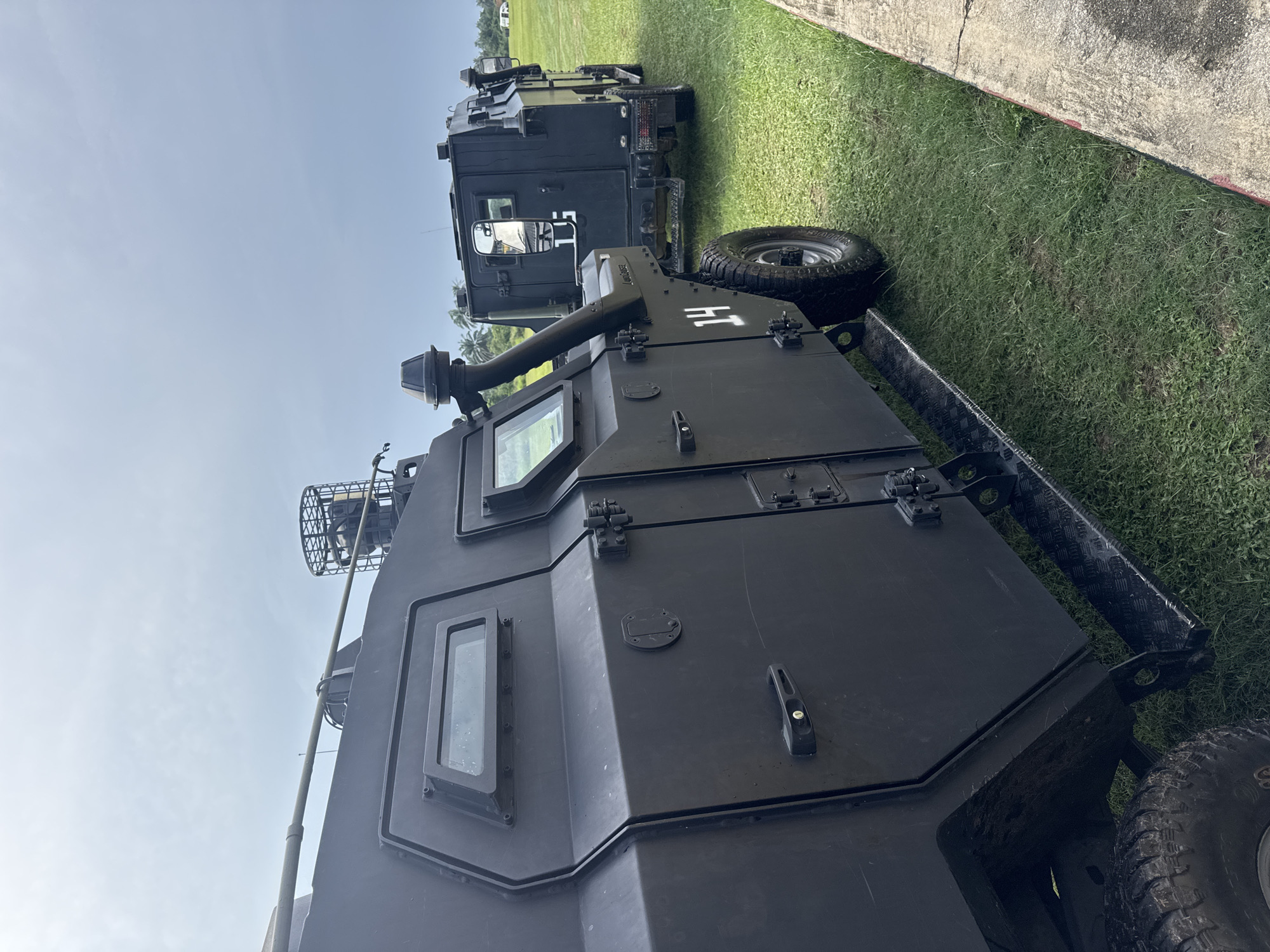 The image shows two black armored vehicles parked on a grassy field, likely in the city of Elele Alimini, Nigeria. The vehicles have visible windows, armored plating, and off-road tires. The closest vehicle has "HT" printed on its side. The field is adjacent to a concrete surface. The sky is visible and clear.