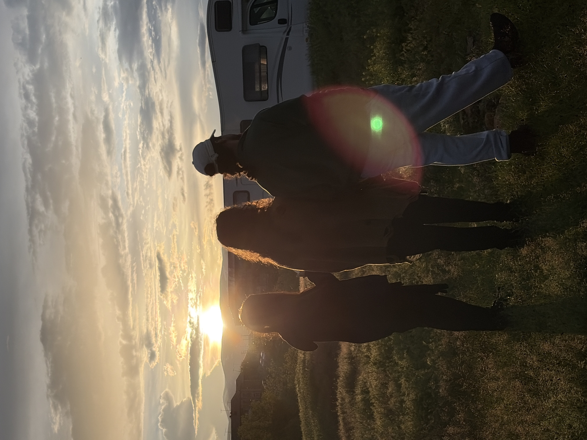 The image shows three people silhouetted against a bright sunset sky. The location appears to be a field of grass with some trees. A recreational vehicle is visible in the background. A man, wearing a baseball cap, appears to be standing behind a woman and a child. The sun is behind the people. The city of Anipemza, Armenia, is not discernible in the image.