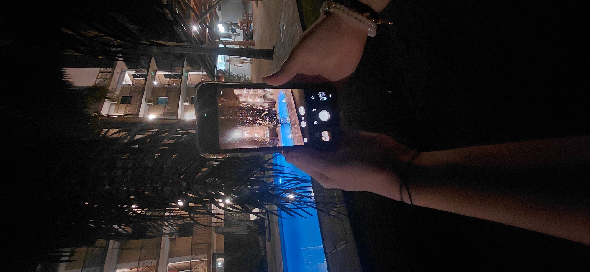 In Presidente Franco, Paraguay, at night, two hands are holding a smartphone, capturing an image of a brightly lit swimming pool and a multi-story building with balconies. The phone displays a reflection of the scene, including palm fronds adorned with small lights. A palm tree is prominent in the foreground, partially obscuring the view. In the background, a man in white clothing walks near a covered outdoor seating area. The environment appears to be a resort or hotel.