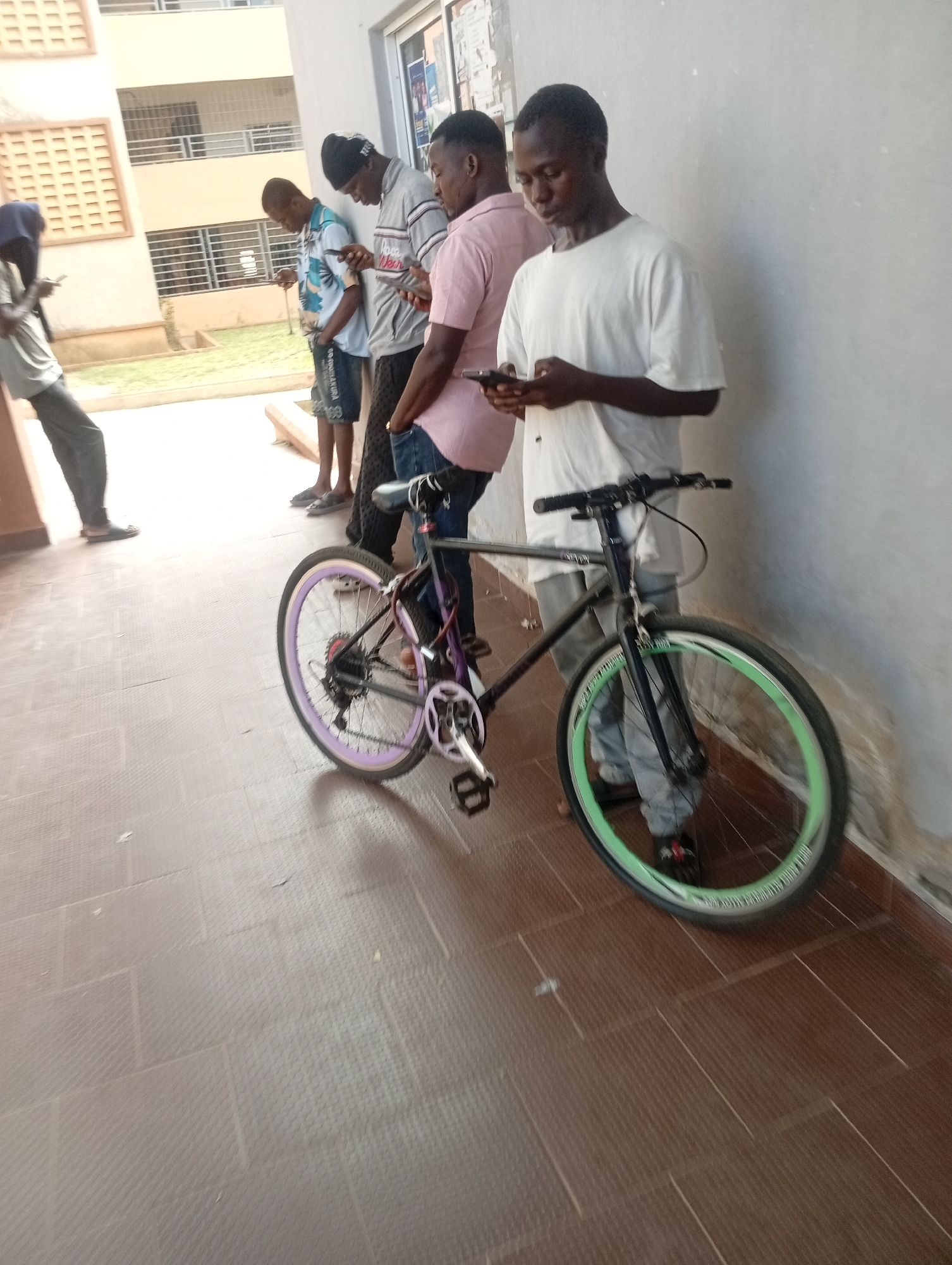 A group of young men are standing around a bicycle, all looking at their phones. The men are in a covered walkway next to a building, possibly a school or dormitory, with a tiled floor. The building has multiple stories and windows, and there is some greenery visible in the background. The men are dressed in casual clothing. One man is wearing a white t-shirt, another a pink shirt, one a grey and white patterned shirt, and another a blue and white patterned shirt. The bicycle is black with purple and green accents on the wheels. The scene appears to be in an urban setting, likely in the city of Jos, Nigeria.