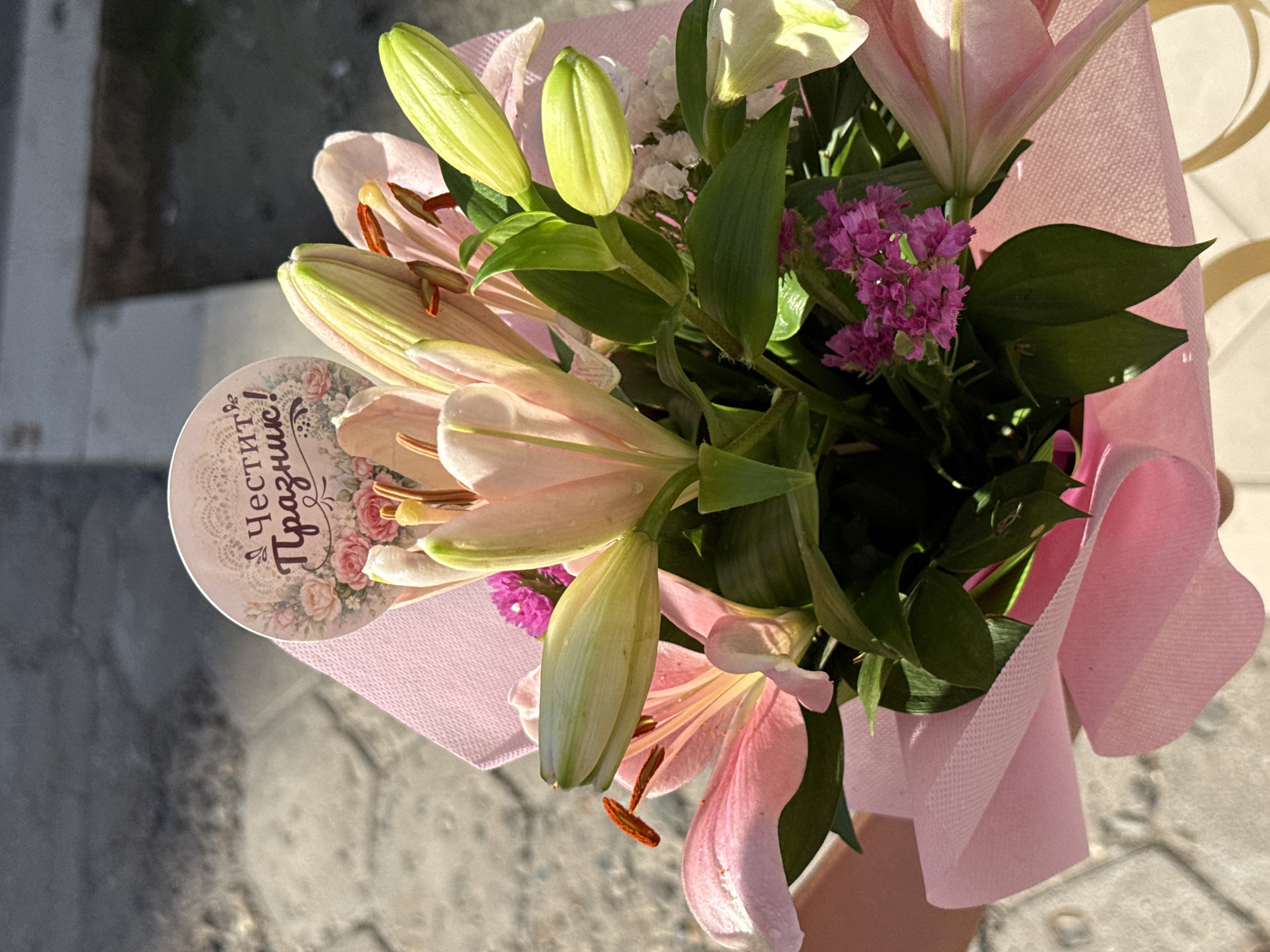 A bouquet of pink lilies and purple flowers wrapped in pink fabric is held in someone's hand. A circular tag with "Честит Празник!" (Happy Holiday!) written on it is attached. The background appears to be a paved surface, likely a sidewalk or pedestrian area. This could be in the city of Pleven, Bulgaria.