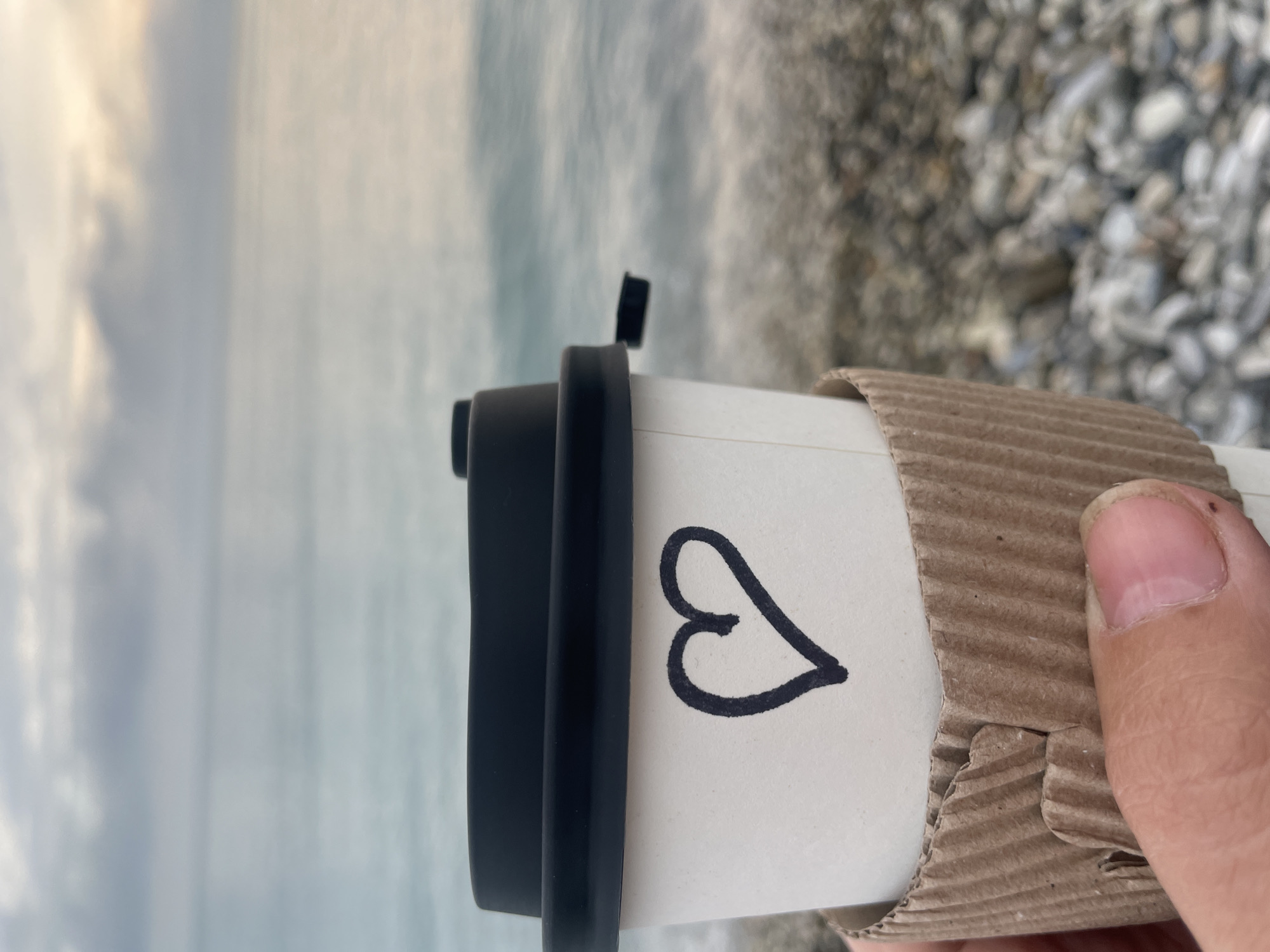 A close-up, horizontally oriented shot depicts a person holding a white paper coffee cup with a black lid and a corrugated cardboard sleeve. A hand grasps the cup's sleeve, with a visible thumb nail. A black heart is drawn on the cup. The background is blurred, but shows a body of water and a shoreline covered with pebbles or small rocks. The location is Olginka, Russia.