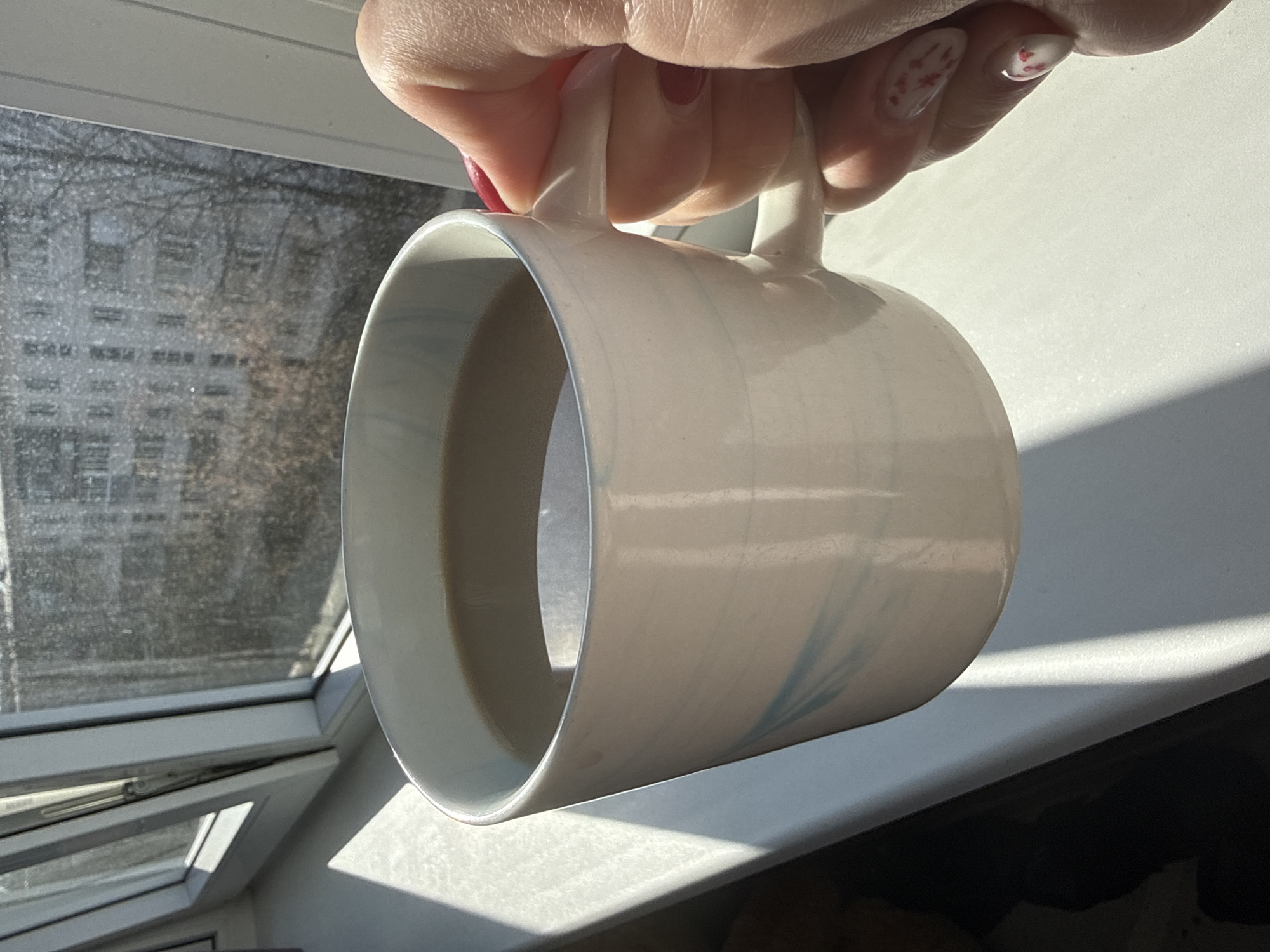 A close-up, high-angle shot shows a hand holding a mug in front of an open window. The mug is white with subtle blue swirls. The interior is light gray, implying that there is some coffee inside. The hand, with red nail polish, is holding the mug by its two handles. The background consists of a building seen through the window, and the windowsill. The image has bright natural lighting. The location is in the city of Chaikovsky, Russia.