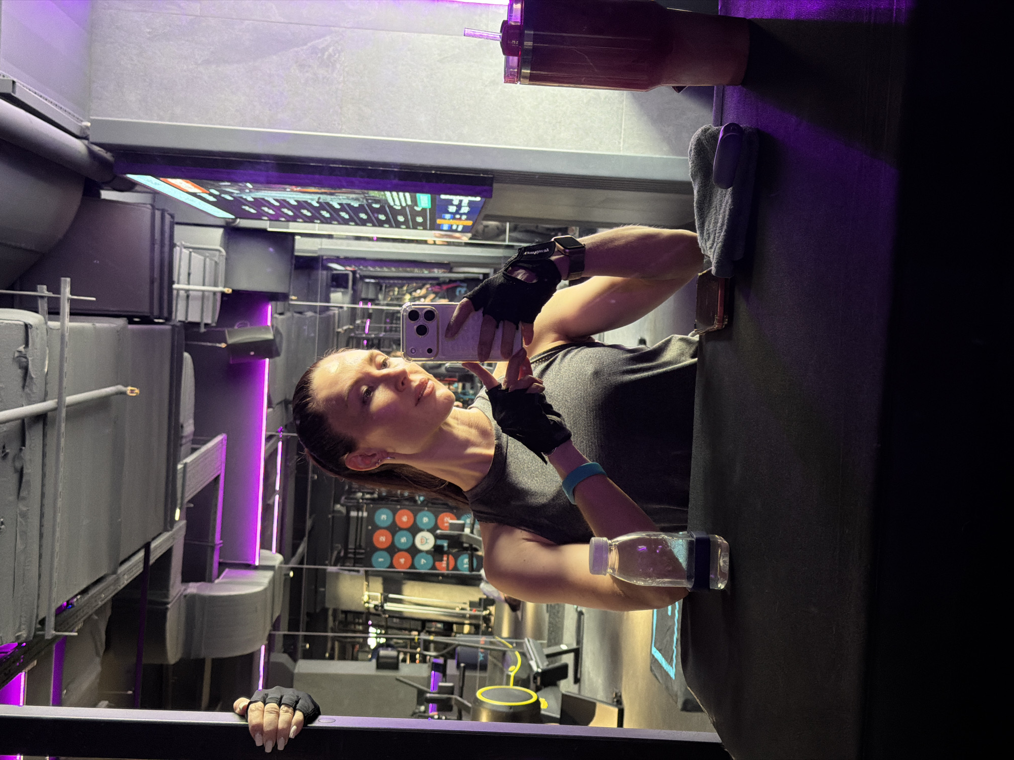 The image features a woman taking a selfie in a modern fitness studio in Perm, Russia. She is positioned slightly to the left, looking into the smartphone held in her left hand, which appears to be a silver-colored iPhone. She has her hair pulled back and is wearing a dark grey athletic top and black fingerless workout gloves; a portion of text, possibly "POWER," is visible on one glove. On her right wrist, she wears a gold-toned smartwatch and a blue fitness band. Her expression is calm and composed.
To her immediate right, a clear plastic water bottle with a blue cap rests on a dark surface. Behind her, a large reddish-purple drink tumbler with a straw and a grey towel are placed on an elevated dark surface.
The background showcases a contemporary gym environment defined by dark walls and ceilings, accented with vibrant purple LED strip lighting. Various pieces of gym equipment are visible, including what appear to be rowing machines or stationary bikes, and other fitness apparatus with metallic components. A wall features a grid of colorful circular targets, possibly for a interactive workout. Several digital screens or displays showing numbers and graphics are integrated into the walls, likely providing workout information. The overall setting is sleek and dynamic, with visible industrial-style ductwork contributing to a modern aesthetic. The woman is engaged in the activity of capturing a moment within this active fitness space.