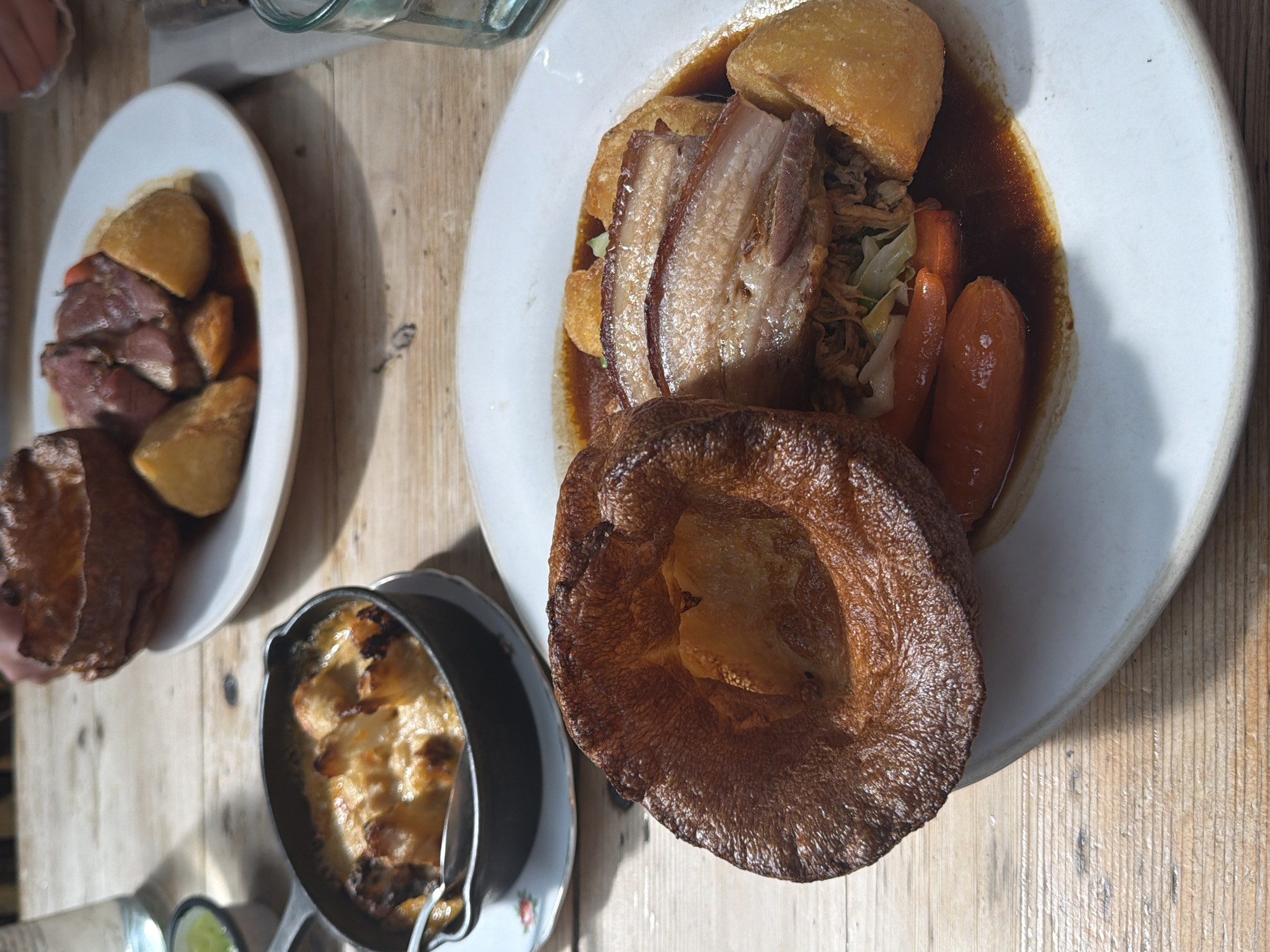 The image captures a culinary scene on a wooden surface, likely a table in a restaurant or dining area. Two plates of food are visible. The plate on the right features a large Yorkshire pudding, sliced pork belly, carrots, and potatoes, all served with gravy. The plate on the left contains slices of roast beef, potatoes, and Yorkshire pudding with gravy. Adjacent to these is a small dish of what appears to be mac and cheese. The setting suggests a traditional British roast dinner. Without additional context, pinpointing the location within the city of London, United Kingdom, is not possible.