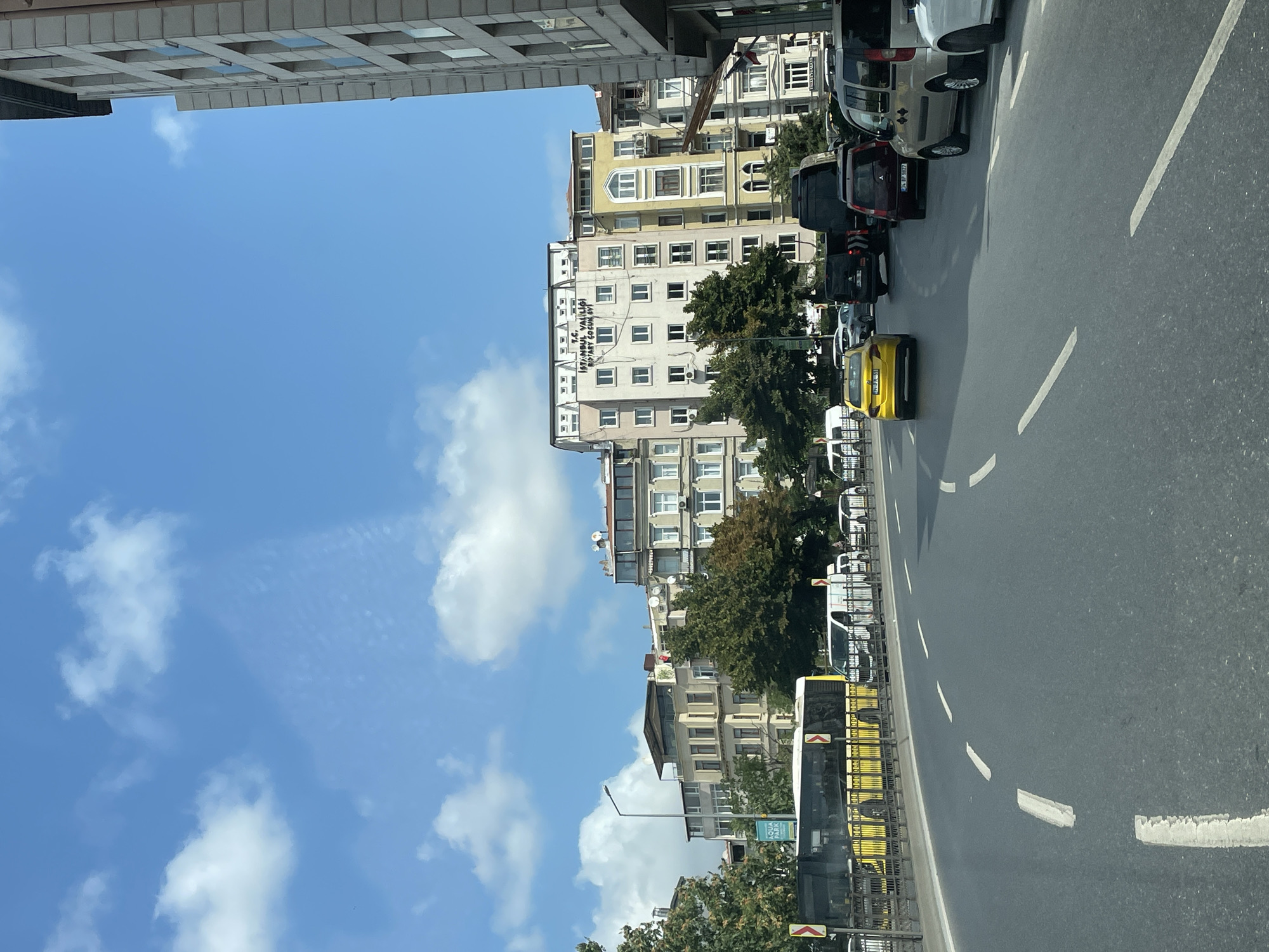 This is a street-level, eye-level, medium shot of a city street in Beyoğlu, Turkey. The scene captures a bright, sunny day with a blue sky dotted with white clouds. The road is asphalt with white lane markings. Several vehicles are present, including a yellow bus on the left, a yellow taxi in the middle lane, and various vans and cars in the right lane. Buildings line the right side of the street, appearing to be multi-story residential or commercial structures with numerous windows. Trees are visible behind the bus and between some of the buildings. The overall impression is of a busy urban environment during daylight hours.