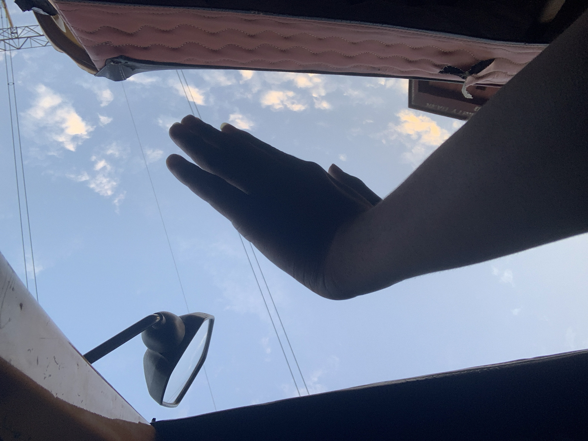 In the city of Obosi, Nigeria, the image captures a low-angle perspective looking upwards towards a bright, partly cloudy sky. A silhouetted arm and hand extend into the frame from the right side, held up towards the sky as if to wave or shield the eyes. In the upper left, a car's side mirror is visible, reflecting the sky. To the right, a pink quilted surface, possibly part of a car's interior or seating, occupies the edge of the frame. A faint outline of power lines stretches across the sky, and a distant structure, possibly a building, can be seen on the horizon. The lighting suggests it is daytime, with the sun casting shadows and illuminating the clouds.