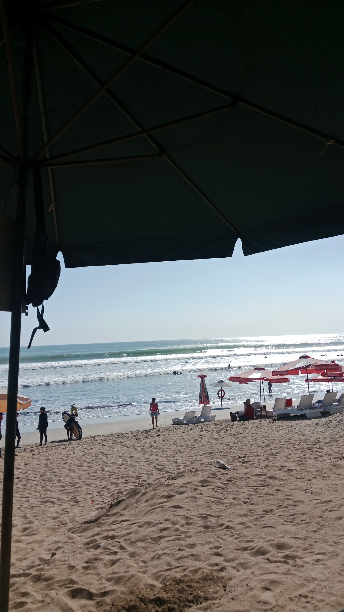 The image captures a sunny day at Kuta Beach, Indonesia, from beneath a large, dark green beach umbrella. The sand in the foreground is textured and appears dry. In the middle ground, several people are visible. To the left, a person in dark clothing stands near the water's edge, and another individual is carrying a surfboard. Further along the beach, a woman in a pink tank top and shorts walks away from the water. A row of white lounge chairs and red umbrellas are set up on the sand. The ocean in the background features gentle waves and several surfers in the water. The sky is clear and bright blue, with the sun reflecting off the water's surface.