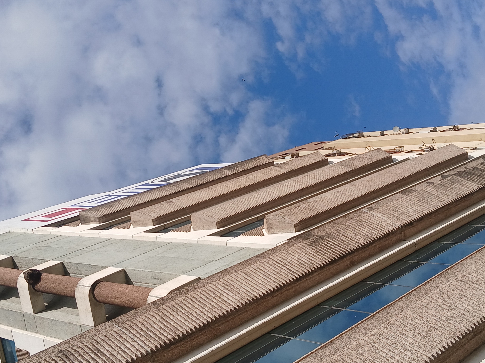 The image depicts a low-angle shot of a building in Ciudad Guayana, Venezuela, against a blue sky with scattered clouds. The building's architectural features include a series of horizontal, textured concrete beams with a repeating saw-tooth pattern along their edges. These beams appear to be structural or decorative elements that run along the building's facade, with rectangular windows reflecting the sky interspersed between them. Additionally, there are cylindrical, pipe-like structures supported by concrete brackets, situated below a section of the building adorned with what appears to be painted red and blue insignia. There are multiple rooftop antennas.