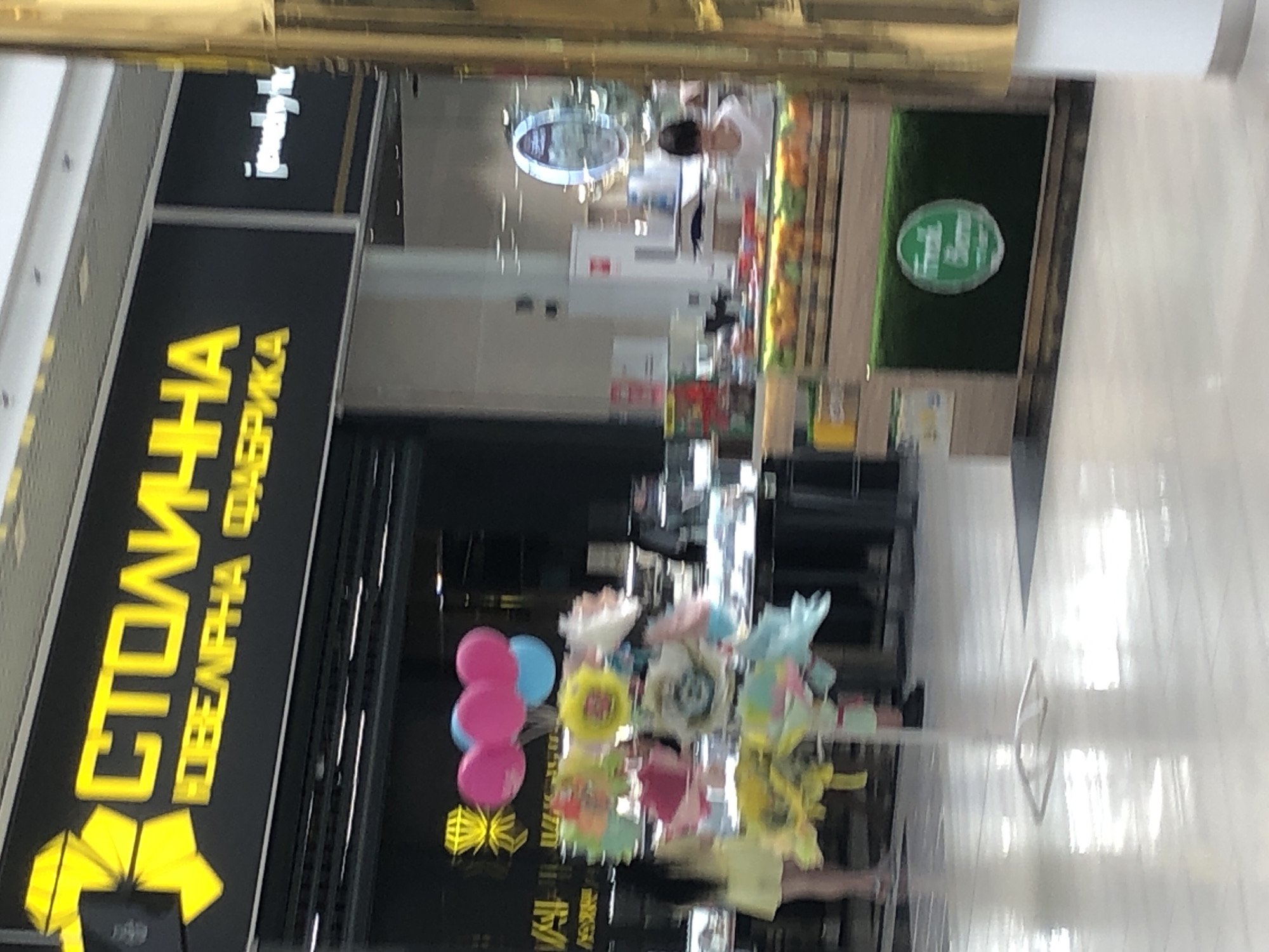 The image captures a brightly lit interior scene, likely a shopping mall, with a focus on a storefront displaying a prominent yellow sign that reads "СТОЛИЧНАЯ ЮВЕЛИРНАЯ ФАБРИКА". To the right of this sign, a smaller white sign with the text "jewelry bx" is visible. In front of the storefront, a display of colorful balloons and flower-like decorations is arranged on stands. A woman in a yellow dress is partially visible on the left, and further to the right, behind a counter stocked with fruits, a person with dark hair is seen. The floor is highly reflective, suggesting a polished surface. The environment appears to be a retail space with various displays and commercial elements.