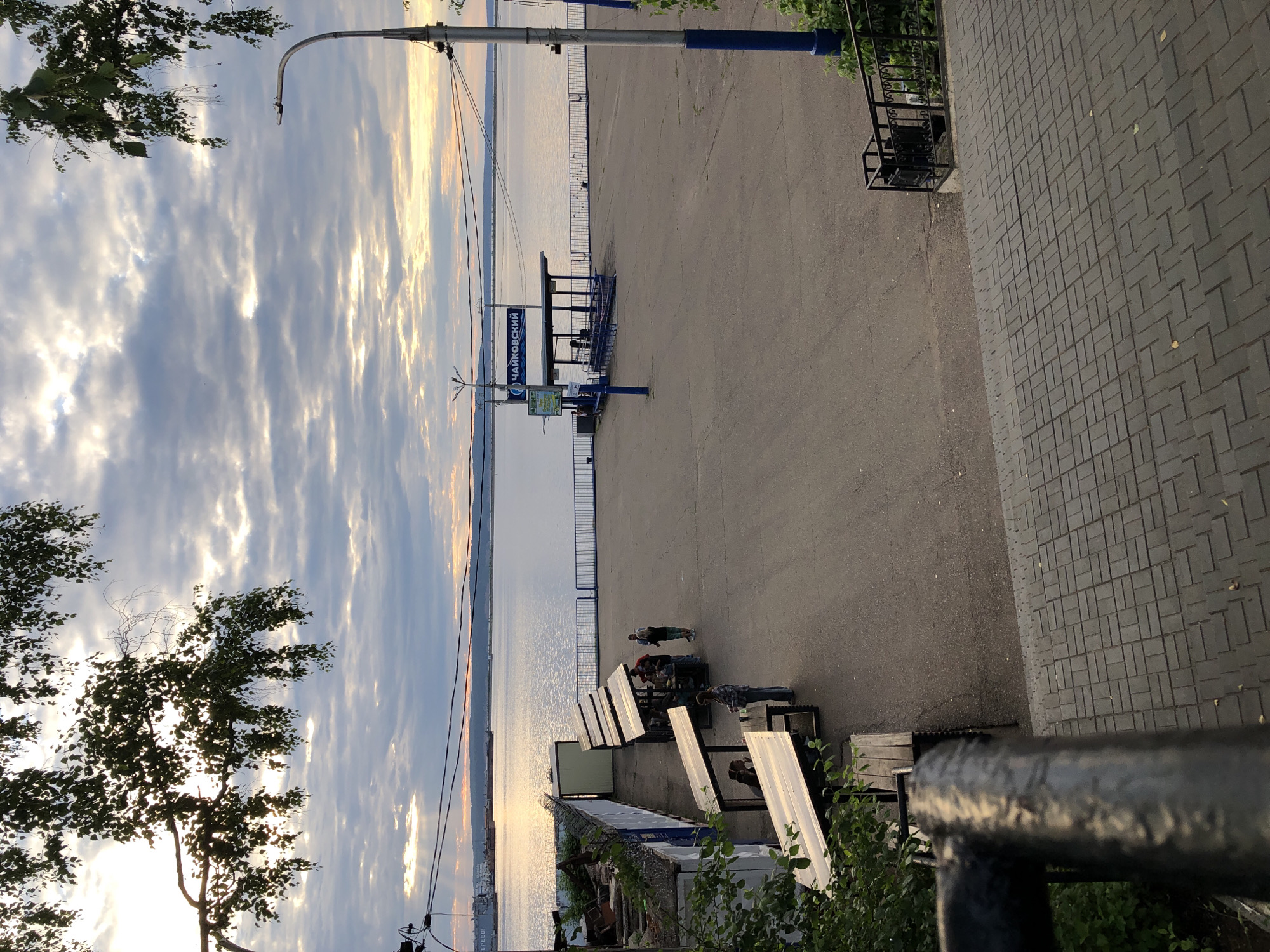 The image depicts a waterfront area in Chaikovsky, Russia, under a cloudy sky with sunlight breaking through. In the foreground, a paved area with brick-like paving stones leads to a dark metal railing. Beyond this, a paved open space features several picnic tables with angled roofs, some occupied by people. A building with a white facade and metal fencing is visible to the left. Further back, a river or large body of water stretches to the horizon, with a distant shoreline and a large ship on the water. A blue and white sign with Russian text, reading "ЧАЙКОВСКИЙ" (Chaikovsky), is mounted on a blue pole next to a covered structure. A streetlamp with a curved arm stands on the right side of the frame. The overall scene suggests a public recreational area by the water.