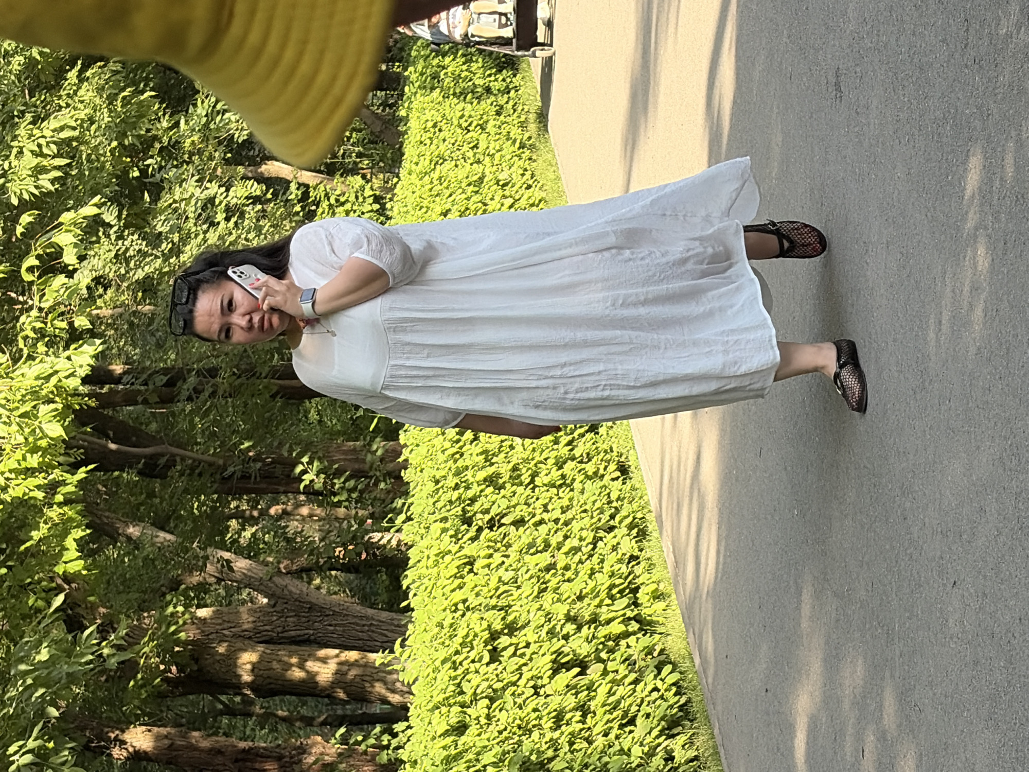 An adult East Asian female walks on a paved path in a park, talking on a silver smartphone. She wears a white, floor-length, loose-fitting dress and black mesh flat shoes. Her hair is pulled back in a ponytail, and she wears sunglasses on top of her head. A bright yellow object, possibly a hat or umbrella, is visible in the upper right corner of the frame, partially obscuring the view. Behind the woman, a vibrant green hedge lines the path, and beyond that, trees with lush foliage create a dappled shade effect on the ground. In the background, a person is visible pushing a stroller, indicating a public park setting. The lighting suggests it is daytime, with sunlight filtering through the trees.