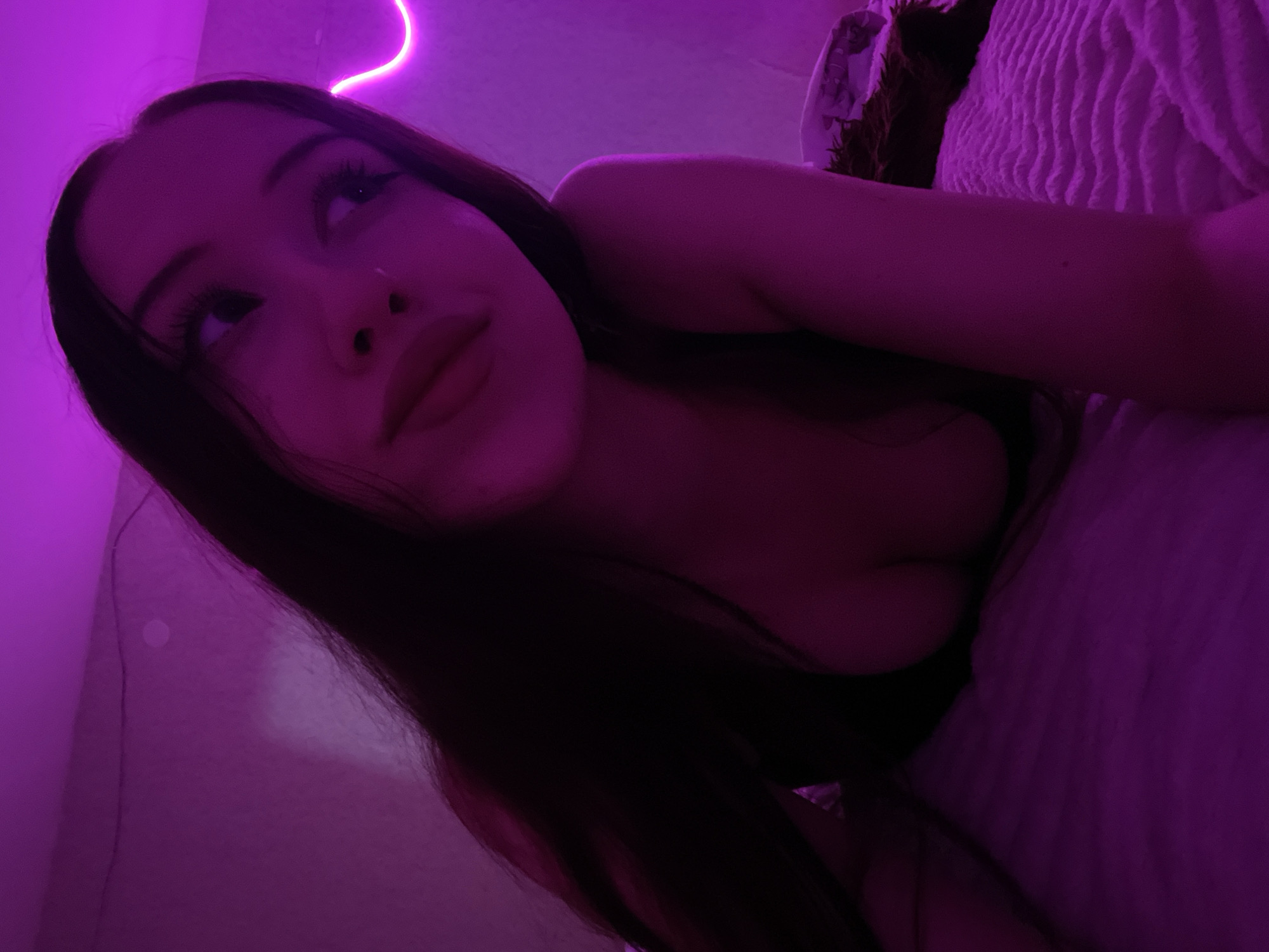 The image shows a young woman lying on a bed in a dimly lit room. The room is illuminated by purple neon lights, which cast a strong glow on the scene. The woman is facing towards the camera, with her head tilted slightly. Her eyes are open and she appears to be looking off to the side. Her hair is long and dark, and it falls around her shoulders. She is wearing a dark-colored top that reveals her décolletage. The bed is covered with a textured pink blanket. In the background, a portion of a wall can be seen, also illuminated by the purple lights. There is no information in the image that can indicate the location in the city of Votkinsk, Russia.
