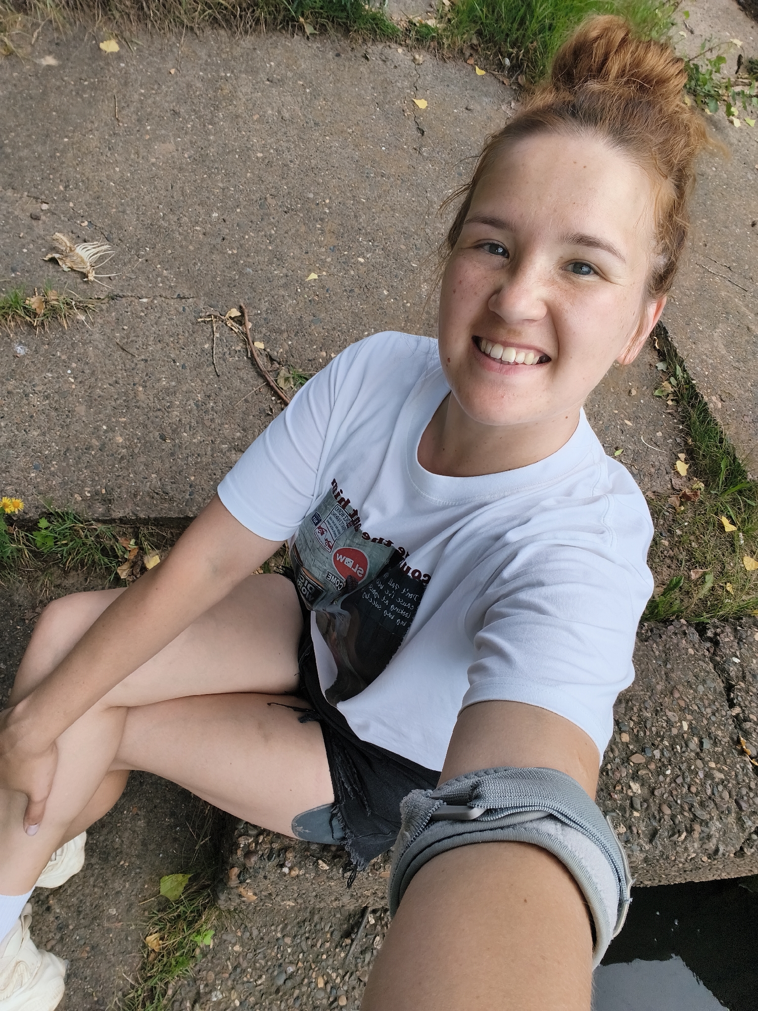 A young woman with reddish-brown hair pulled into a messy bun smiles at the camera, holding a phone in her right hand to take a selfie. She is wearing a white t-shirt with graphic text and images, black ripped shorts, white socks, and light-colored sneakers. She is sitting on a cracked concrete surface with patches of grass and fallen leaves around her. Her legs are crossed, and her left arm rests on her bent knee. A gray wristband with a zipper is visible on her right forearm. The scene appears to be outdoors, possibly a park or pathway, given the concrete and grassy elements. The lighting suggests it is daytime, with natural light illuminating the scene. The woman's expression conveys happiness and a relaxed demeanor. The setting suggests a casual, everyday moment. The location context provided is Chaikovsky, Russia.