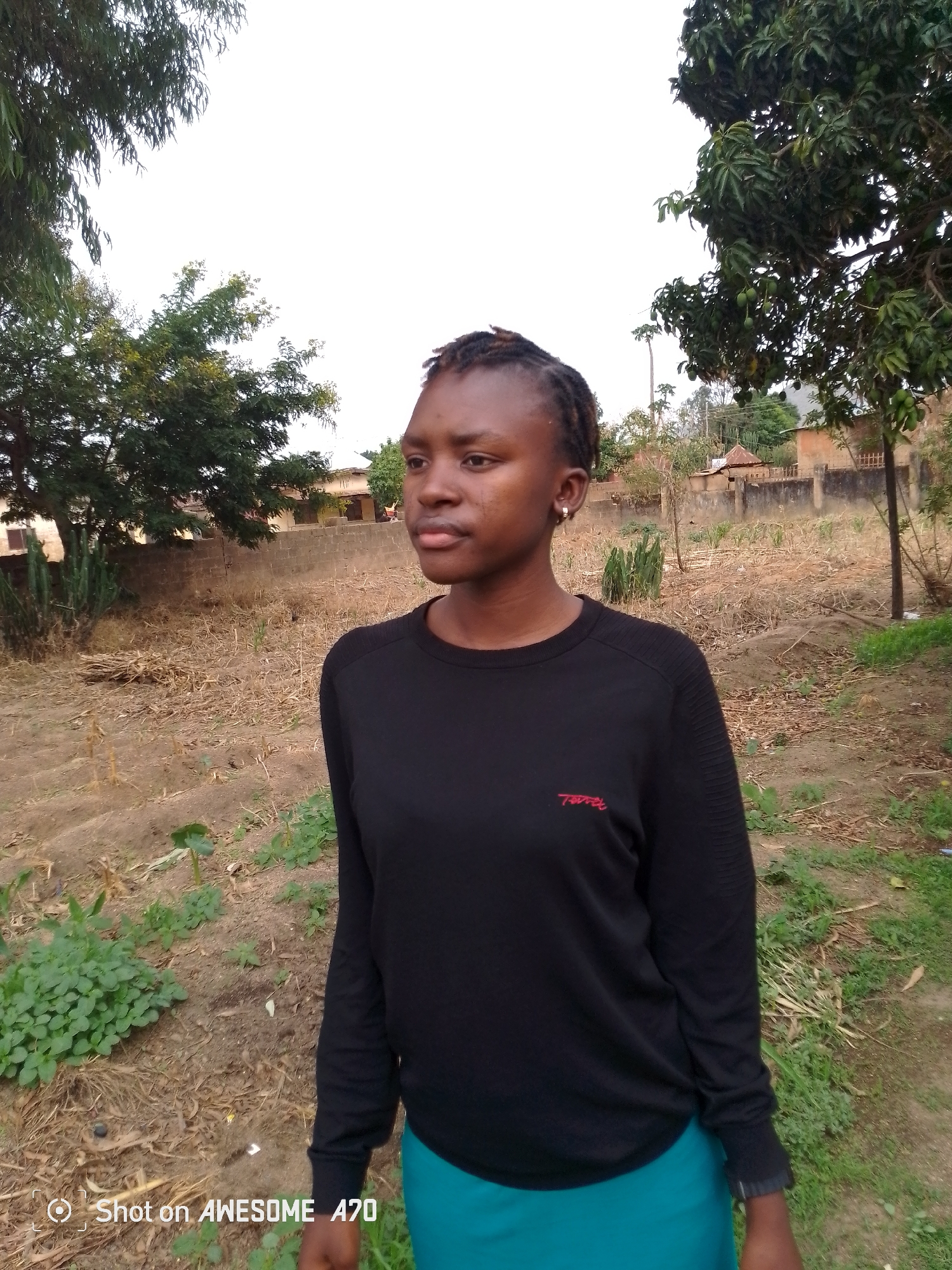 A young woman stands outdoors in an area that appears to be a yard or field in Unknown, Unknown. She has dark hair styled with small braids or twists, and wears a dark long-sleeved shirt and a teal skirt. Her gaze is directed slightly to the left. The background includes trees, a fence, and buildings in the distance. The ground has patches of dry vegetation and some green plants. The image is labeled in the bottom left corner with "Shot on AWESOME A70".