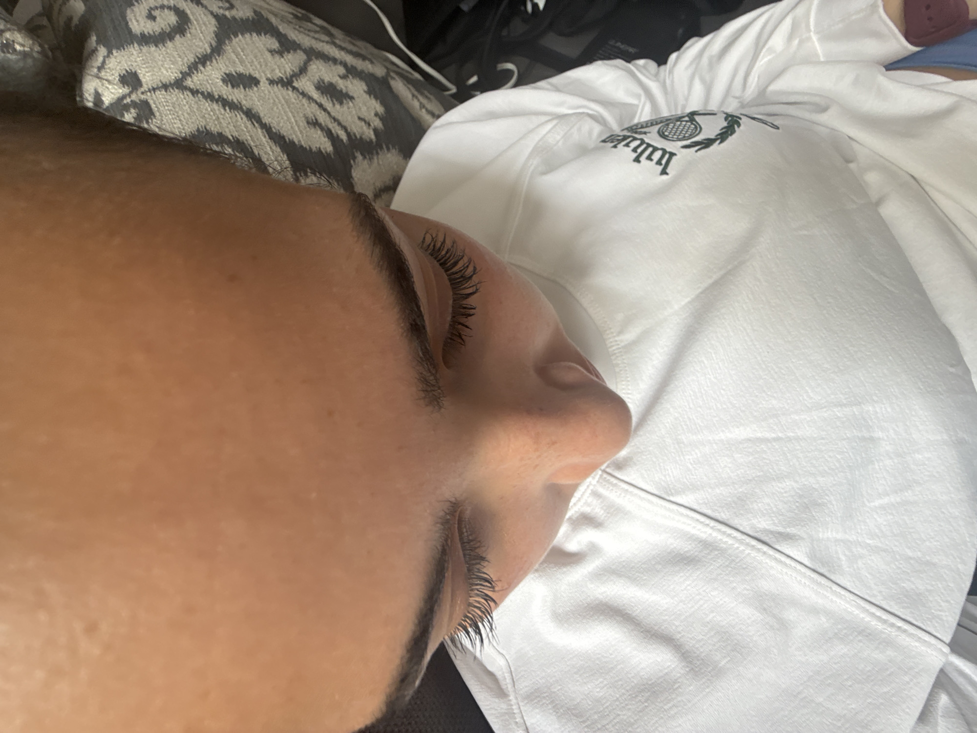 A person's face is shown in close-up profile, their eyes closed. The person is lying on a patterned cushion and wearing a white sweatshirt with an emblem on the front. The location is not discernible from the image. No actions or interactions are visible.
