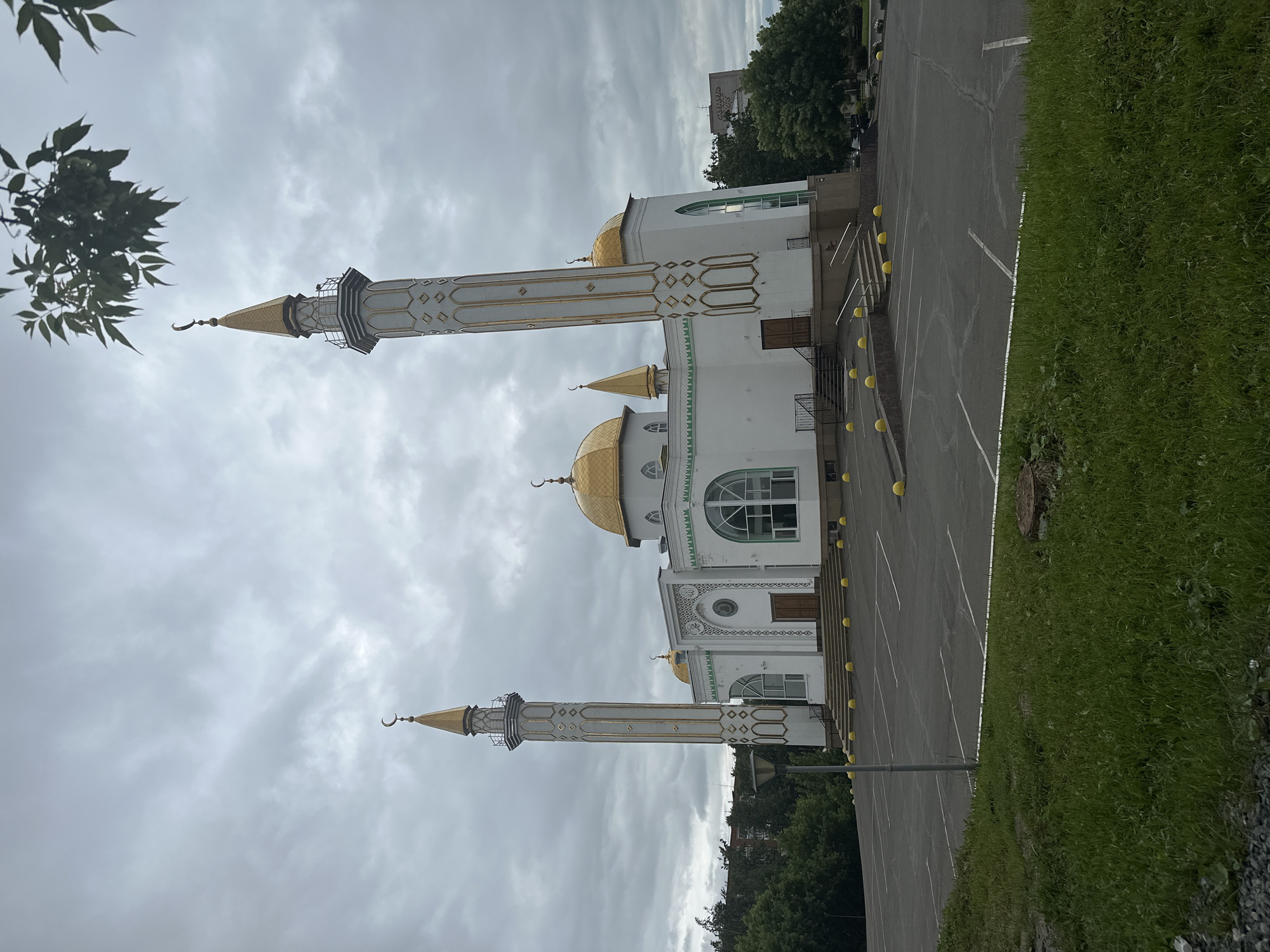 The image displays the Iman Mosque in Izhevsk, Russia, under an overcast sky. The mosque is a prominent white structure featuring multiple golden domes and two tall, slender minarets. The central dome is the largest, flanked by smaller golden domes, all topped with golden spires bearing crescent moon finials. The two identical minarets are white with ornate vertical golden decorative elements and diamond patterns, each capped with a golden spire and crescent moon. The main facade of the mosque exhibits arched windows, intricate gold-colored patterns around a central wooden double door, and subtle green trim along rooflines and window frames.
Multiple sets of dark steps with silver handrails lead up to the mosque's entrances. Numerous bright yellow spherical bollards line the base of the building and the perimeter of an adjacent asphalt parking lot. The parking lot, marked with white lines, appears empty. In the immediate foreground, green grass covers a sloped area. Beyond the mosque, a dense arrangement of green deciduous trees is visible, partially obscuring other distant urban or suburban buildings. One background building has partially visible Cyrillic script ("АЛЛАН-"). A branch with green leaves extends into the upper right corner of the frame. The sky is predominantly grey with visible cloud formations, indicating an overcast daytime setting. No people are present in the scene.
