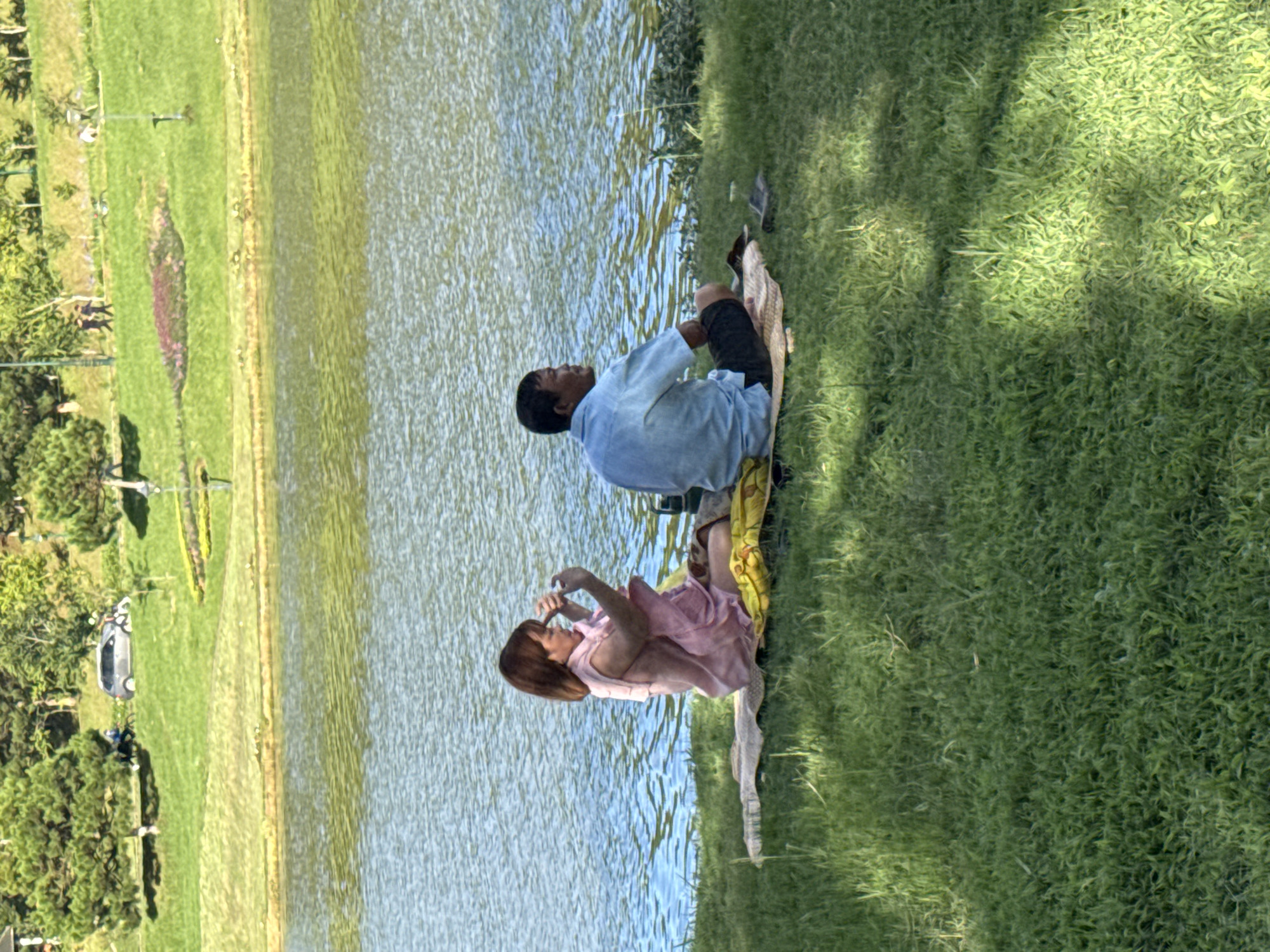 The image depicts a serene lakeside scene in Phường Xuân Hương - Đà Lạt, Vietnam. In the foreground, two individuals are seated on a mat on the grass, facing a body of water. A woman, on the left, is wearing a pink dress and appears to be looking at her phone. A man, on the right, is wearing a light blue shirt and dark shorts, looking away from the camera towards the water. The background reveals a lush green hillside with scattered trees and a few distant figures walking. A road with a car and a cyclist is visible further up the hill. The lake's surface is rippled, reflecting the surrounding greenery and the sky. The overall atmosphere is peaceful and recreational.
