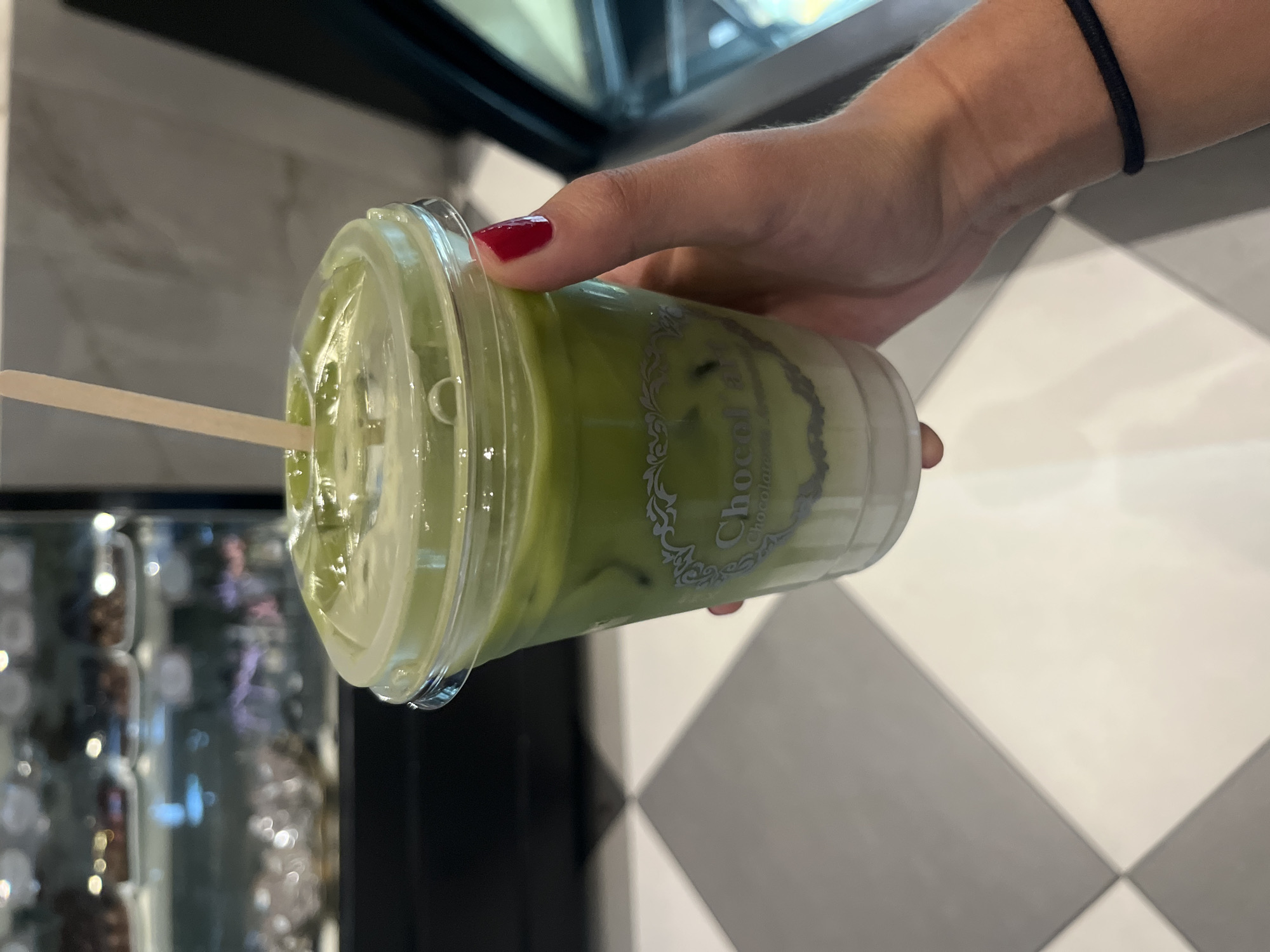 A hand with red nail polish is holding a plastic cup of green matcha latte. The cup has a clear lid and a straw. The label on the cup is partially visible and it says "Chocolat D'Amour." The background shows a black and white checkered floor and a glass display case. The image appears to be taken indoors at a coffee shop or café in the city of Puebla, Mexico, based on the branding on the cup.
