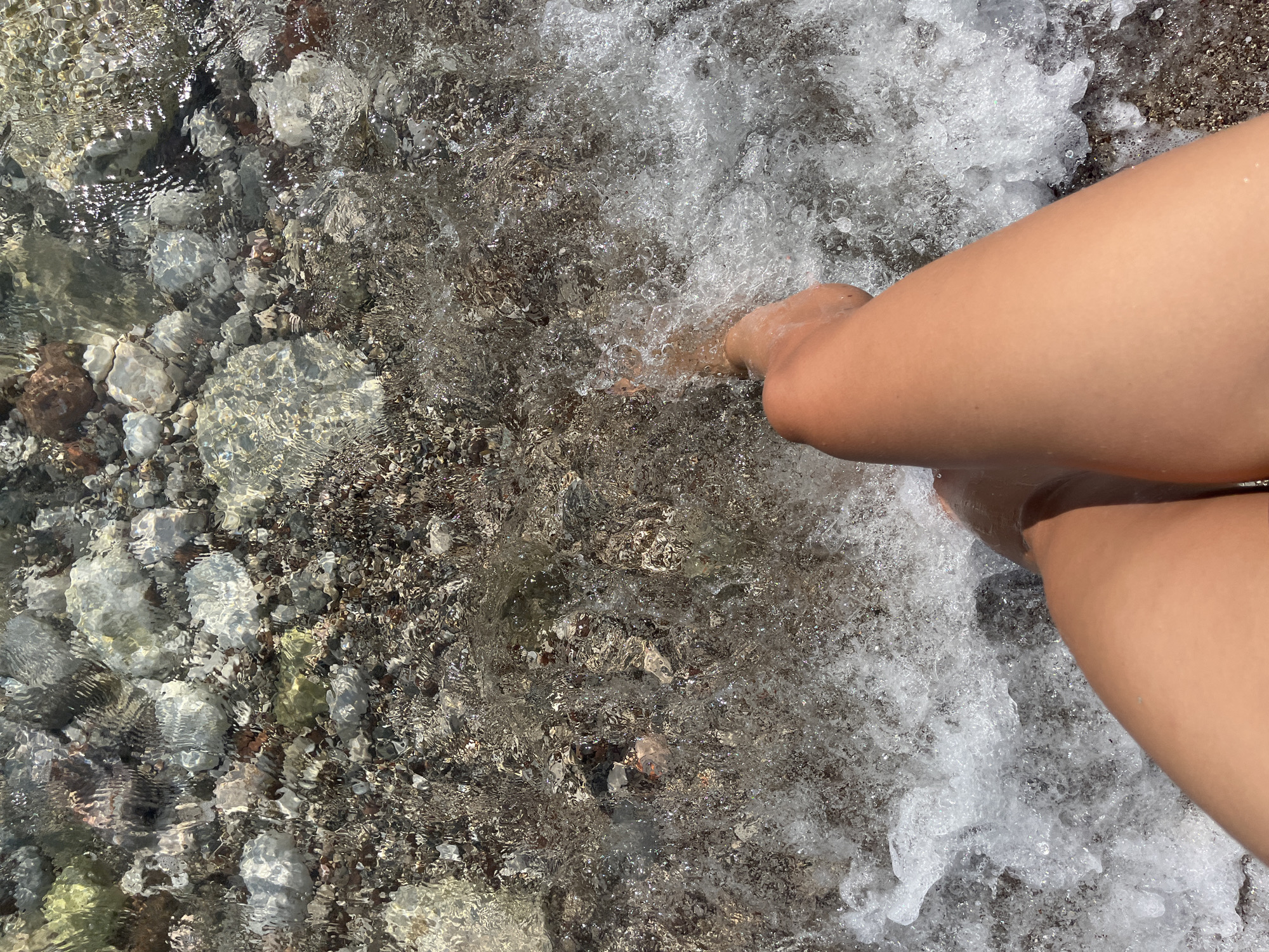 The image shows a person's legs and feet partially submerged in clear, shallow water. The water is moving, creating ripples and bubbles. Beneath the water, a bed of small, smooth pebbles is visible, with some larger, lighter-colored rocks scattered among them. The light refracts through the water, creating shimmering patterns on the pebbles. The person's skin tone is warm and tanned, suggesting they are outdoors in sunlight. The scene evokes a sense of relaxation and enjoyment of a warm, sunny day, likely at a beach or a waterfront location. The pebbles and clear water suggest a coastal environment, and the provided location context of Bar, Montenegro, reinforces this interpretation. The time of day appears to be daytime, with bright lighting conditions evident from the reflections on the water. There is no visible text in the image.