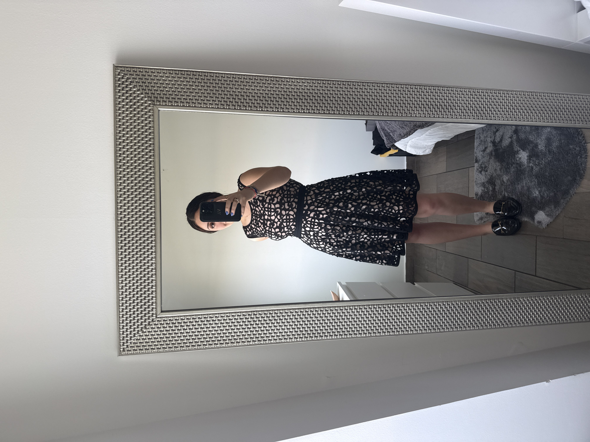A female subject is depicted taking a mirror selfie in an interior room, located in Mexico City, Mexico. The subject, visible from head to mid-calf in the mirror's reflection, wears a sleeveless black dress featuring a laser-cut pattern that reveals a light-colored underlay, with a solid black band at the waist. She wears black patent leather loafers with gold-toned buckle details. Her right hand holds a dark-colored smartphone, positioned to obscure the upper portion of her face. Light blue nail polish is visible on her fingers, and multiple bracelets are worn on her left wrist. The rectangular, full-length mirror possesses a textured, woven metallic frame. The reflected background includes plain white walls. To the left of the subject, a white multi-drawer dresser is visible, topped with a small wooden bowl. To her right, a segment of a bed with white linen, a grey throw blanket, and yellow fabric is present. The floor is composed of dark wood or tile, with a grey faux fur rug placed in front of the mirror.