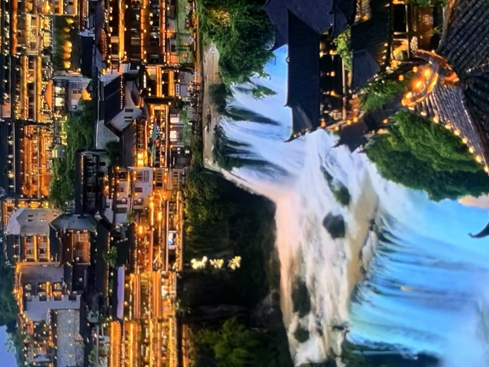The image depicts a nighttime view of a scenic waterfall cascading down a cliff face. Several multi-story buildings with glowing lights are visible on the hillside above and around the waterfall, suggesting a town or city integrated with the natural landscape. The architecture of the buildings appears to be traditional, with dark tiled roofs and many illuminated windows and decorative lights. Some of the buildings are situated on terraces carved into the hillside. In the foreground, a traditional dark-tiled roof structure with decorative eaves and ornate lighting is prominent. Lush green vegetation covers parts of the cliff face and the surrounding areas. The waterfall itself is wide and powerful, with turbulent white water at its base, illuminated by the lights from the town and potentially some artificial lighting in the water. There are no discernible people or specific actions captured in the scene. The image does not show any features related to the city of Stavropol, Russia.