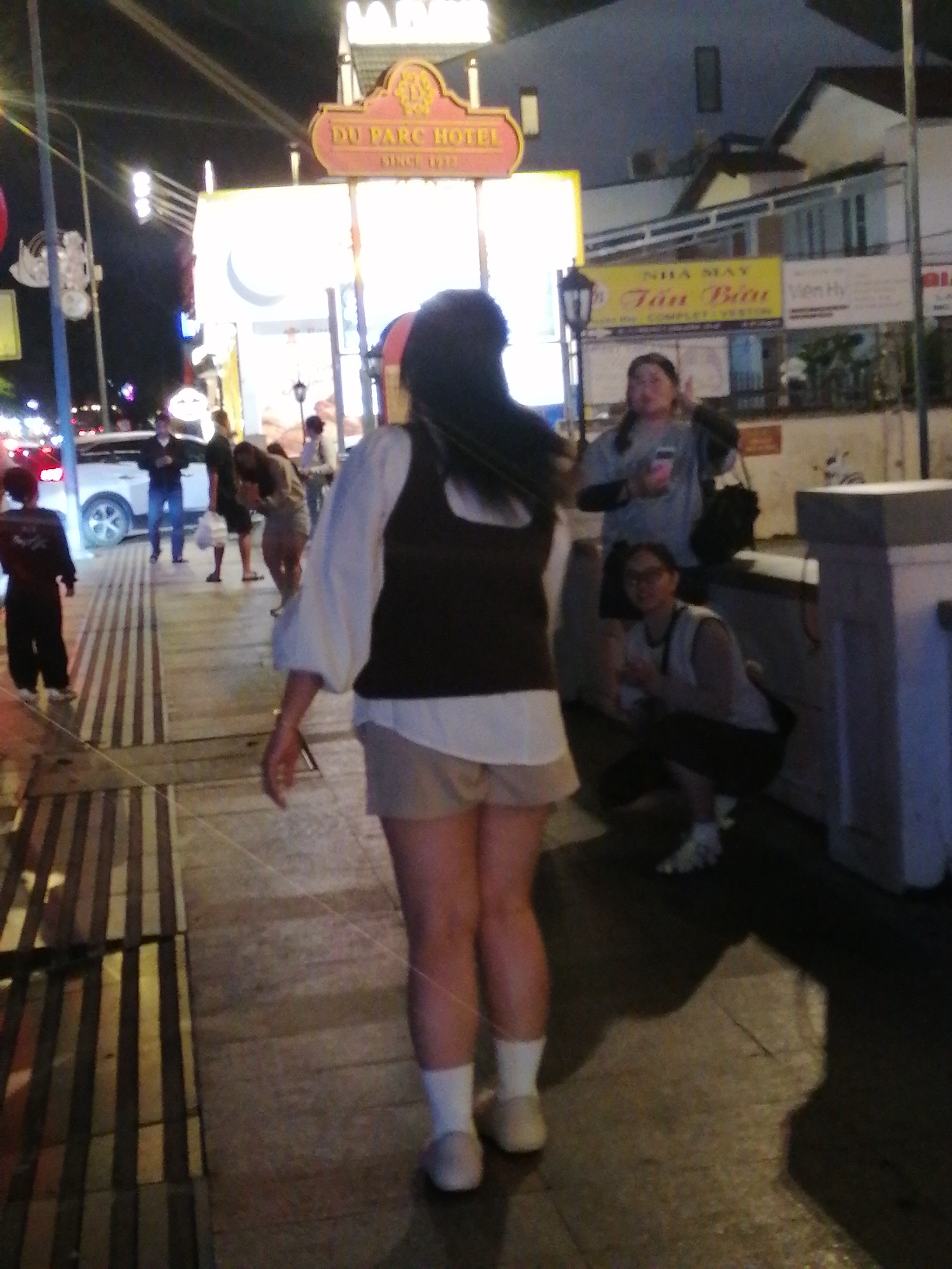 The image captures a nighttime street scene in Da Lat, Vietnam, featuring the facade of the DU PARC HOTEL, illuminated by prominent signage. A woman, viewed from behind, is the central figure, wearing a white shirt with a dark vest and light-colored shorts. Her gait suggests she is walking away from the camera. In the background, other individuals are present: a woman in a gray shirt is standing, another woman is crouched, and a child is visible in the distance to the left. Vehicles, including a white car, are also present on the street. The environment is characterized by streetlights and building lights, typical of an urban setting at night.