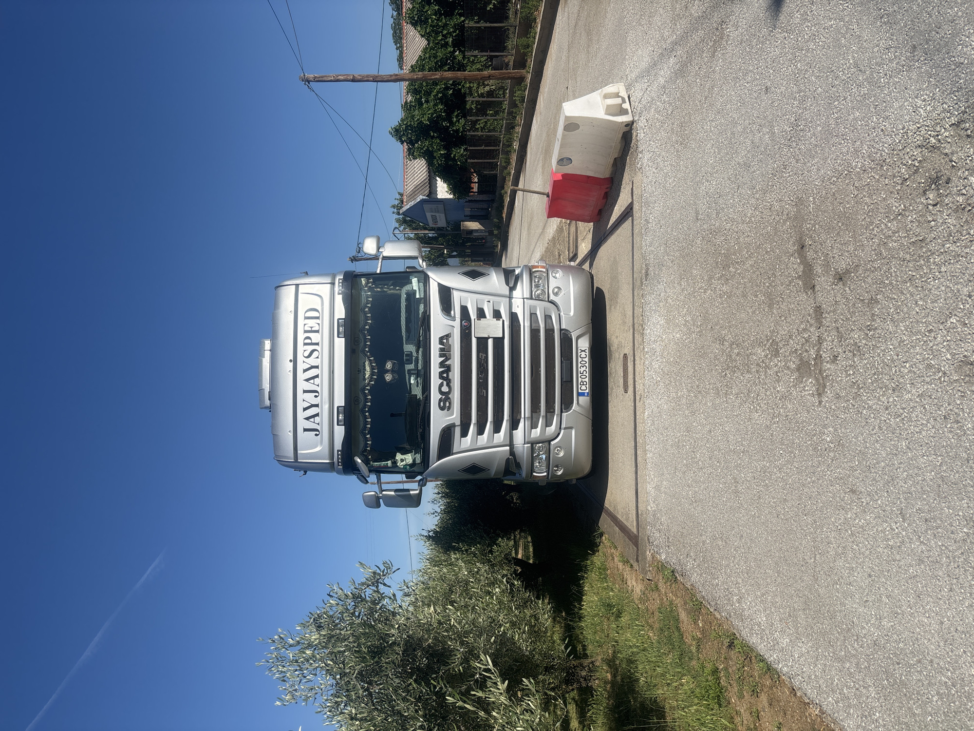 A silver Scania truck with the company name "JAYJAYSPED" is parked on a weighbridge. The truck has a Bulgarian license plate, CB 0530 CX. To the right of the truck are two temporary barriers, one red and one white. In the background, there are buildings with red roofs, trees, and a wooden utility pole. The scene is outdoors, under a clear blue sky with a faint contrail. The location is identified as Kikidio, Greece.