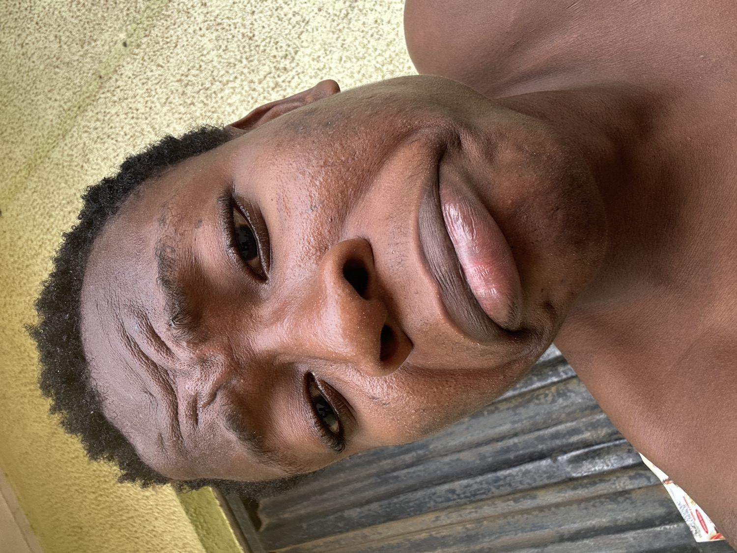 A close-up, slightly angled shot shows a Black man's face. His complexion is dark brown, with some shine on his skin. He has short, dark hair and visible forehead wrinkles. His lips are pursed. The background features a yellow textured wall on one side and a gray corrugated surface on the other. The location is unknown.