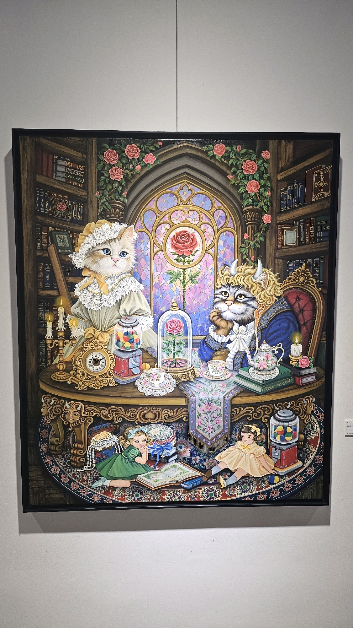 The image depicts a framed oil painting displayed on a white wall, likely in an art gallery or exhibition space. The painting itself is a detailed, fantastical scene that reimagines the story of Beauty and the Beast with anthropomorphic cats as the characters.
In the center of the painting, two cats are seated at a table. The cat on the left, dressed in a Victorian-style yellow dress and bonnet with lace trim, has bright blue eyes and light-colored fur. Beside her is a vintage-style gumball machine filled with colorful candies and a gold-trimmed clock. A glass dome covers a single red rose, a symbolic element from the original story.
The cat on the right, depicted as the Beast, has a more rugged appearance with brown fur and small horns on its head. It is wearing a blue coat with gold trim and a white cravat. A teapot adorned with pink roses and teacups sit on a stack of books, one of which is titled "Once Upon a Cat."
In the background, a large, ornate stained-glass window with a rose motif is visible, framed by Gothic-style architecture and ivy with pink roses. Bookshelves filled with antique-looking books line the walls on either side of the window.
In the foreground, two young girls are depicted as dolls on a richly patterned rug. The girl on the left wears a green dress and is leaning on her elbow, looking at an open book. The girl on the right is wearing a yellow dress and sitting with her legs extended.
The overall style of the painting is whimsical and illustrative, with vibrant colors and intricate details. The lighting in the gallery is soft and even, highlighting the artwork. The image is taken from a medium distance, capturing the entire painting and its immediate surroundings. The white wall provides a neutral backdrop that emphasizes the artwork.
This artwork is located at the Bangkok Art and Culture Centre (BACC), a prominent cultural venue in Bangkok, Thailand, known for hosting contemporary art exhibitions. The exhibition shown in the image features the unique "Beauty and the Beast" cat-themed artwork by an artist named "Once Upon a Cat," whose name is visible on the book within the painting.