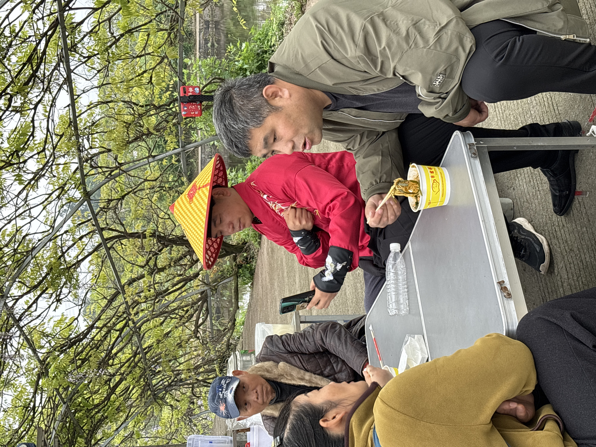 At an outdoor table near a body of water in Xianfeng, China, four adults are gathered. One man, wearing a traditional yellow conical hat and red jacket with crane embroidery on the sleeves, eats noodles from a yellow instant noodle cup with chopsticks. A second man, in a khaki jacket, leans in, looking at the noodles. A third person wearing a brown jacket and baseball cap looks toward a fourth individual in a yellow shirt and grey pants. A plastic water bottle sits on the table beside the noodles. A phone is also on the table. Bare trees frame the background.