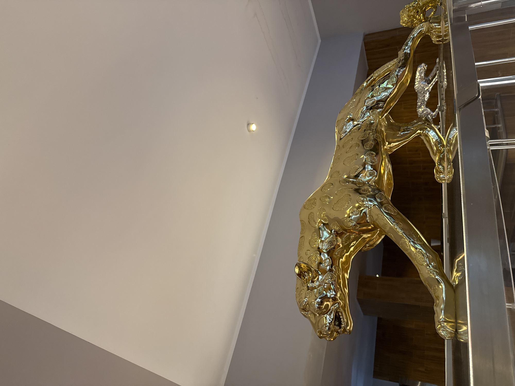 This image depicts a large, golden sculpture of a leopard in motion, placed on a reflective surface. The sculpture is highly detailed, with textured spots and an open mouth. A smaller, silver leopard sculpture is positioned underneath the larger one, also in a stalking pose. The background features wooden paneling and a plain gray wall, leading up to a white ceiling with a recessed light fixture. The overall impression is one of opulence and a sophisticated interior design. The exact location in Nordhausen, Germany, is not discernible from the image itself, but the setting suggests an indoor, possibly public or residential space.