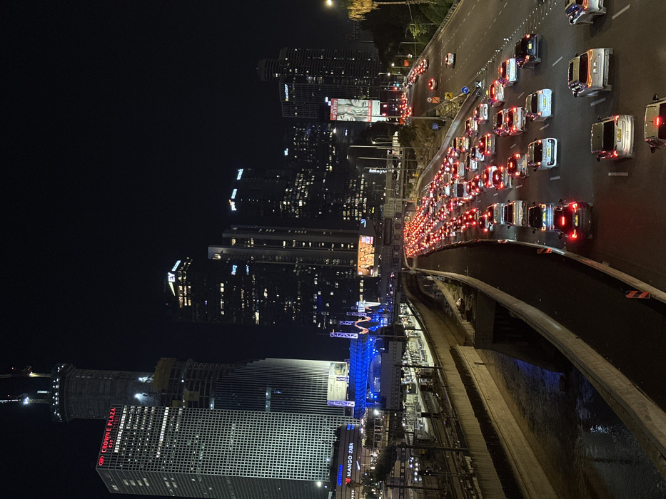 A night cityscape features dense traffic on a multi-lane highway illuminated by car headlights. Tall, modern buildings with numerous lighted windows dominate the skyline on the left. A prominent structure displays the "Crowne Plaza" logo. Construction scaffolding is visible on one building. The scene includes signage for "Mango Zara." Blue lights highlight an event space or structure below the buildings. Overall, the image captures the urban hustle and bustle at night.