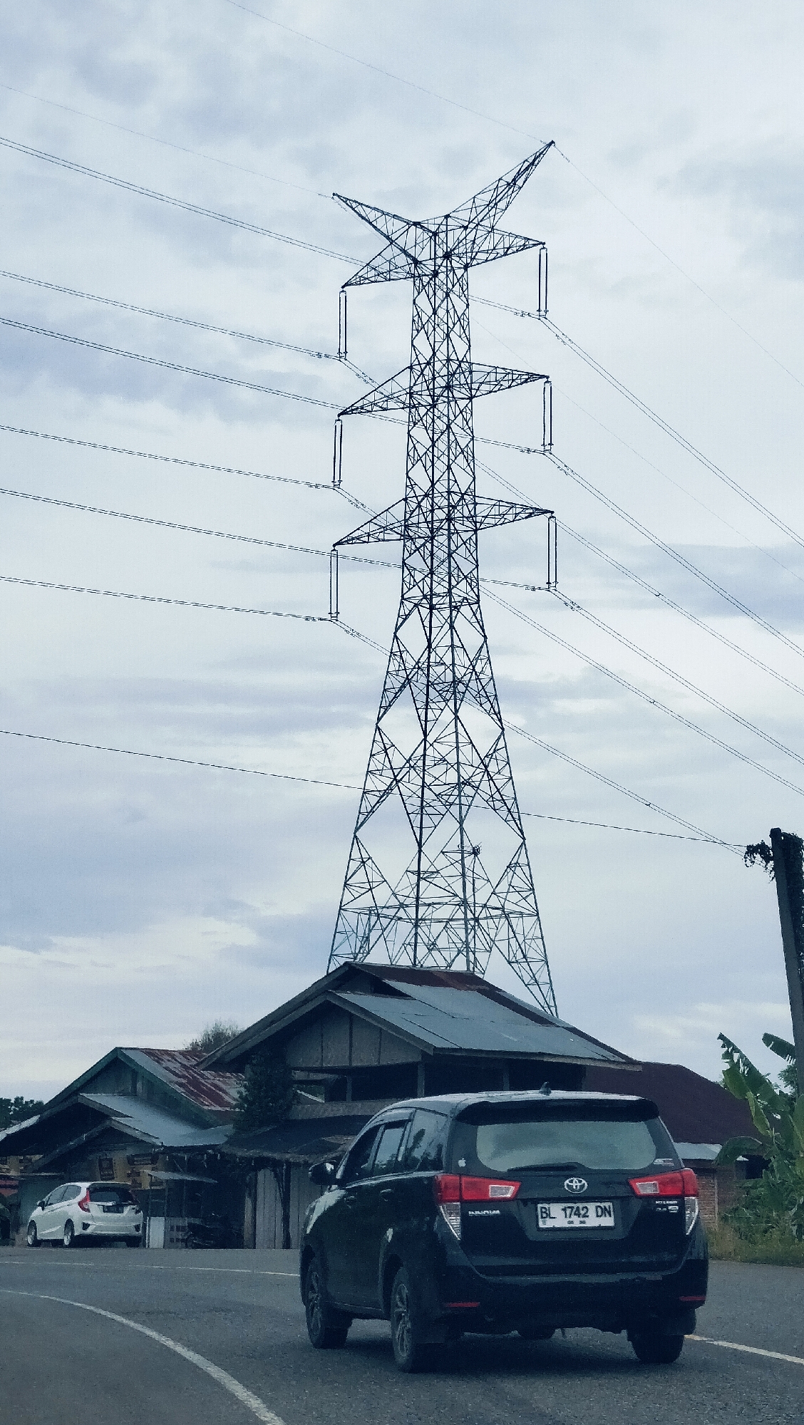 This image captures a black Toyota Innova driving on a paved road, viewed from its rear-right, against a backdrop dominated by a large electricity transmission tower and an overcast sky. The setting appears to be a semi-urban or rural area in Lambarotunong, Indonesia.
The towering power pylon, with its intricate metal lattice structure and multiple overhead power lines, stands prominently in the upper half of the frame. Below the pylon and behind the black car, several weathered buildings with corrugated metal roofs are visible, suggesting local architecture. Another white car, possibly a Honda Jazz/Fit, is parked further down the road on the left side.
The sky is cloudy, indicating an overcast day, perhaps in the morning or late afternoon, with soft, diffused lighting. The road features a white broken line along its curve. The black Toyota Innova has a clear license plate, "BL 1742 DN," with "BL" being a regional code for Aceh, Indonesia. The car's make "TOYOTA" and model "INNOVA" are also visible on its rear. Sparse vegetation, including banana leaves, can be seen on the right side of the frame. The scene portrays a typical day, focusing on infrastructure and local transportation.