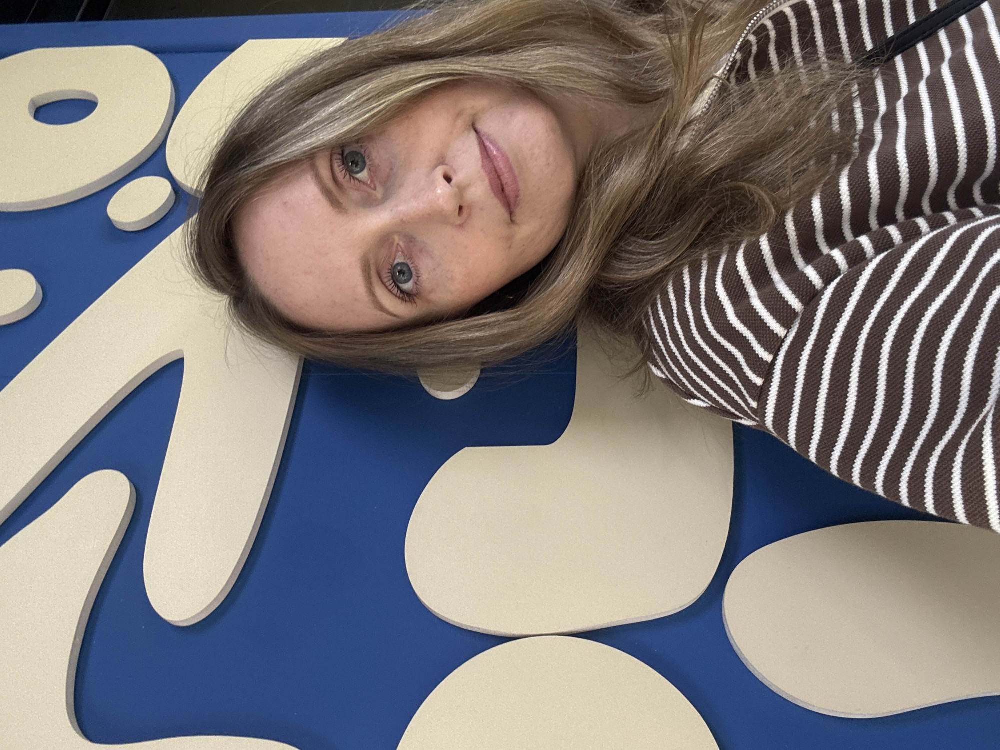 A young woman lies on a blue surface decorated with off-white shapes. Her blonde hair is parted, and she wears a brown and white striped zippered sweater. The location is not identifiable as Votkinsk, Russia.