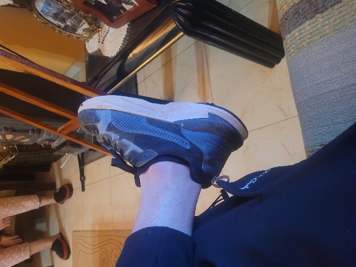 An indoor scene in Κωστήτσι, Greece, depicts a person's lower left leg and foot in the immediate foreground, positioned over a patterned sofa or chair. The individual wears dark blue or black sweatpants with a white logo on the thigh and a dark grey/black athletic shoe with a tan sole. The floor is tiled with light-colored square tiles, partially covered by a light-patterned rug. In the background, two pairs of bare feet wearing red and black slip-on slippers are visible on the floor, suggesting the presence of other individuals. To the right, a dark, ornate structure, possibly a stove or fireplace, stands beside a wooden frame leaning against a warm yellow wall. A dark, reflective table, adorned with a white crocheted doily and two framed photographs (one gold, one reddish-brown), is positioned against the wall. A dark, ribbed decorative object is visible below the table.