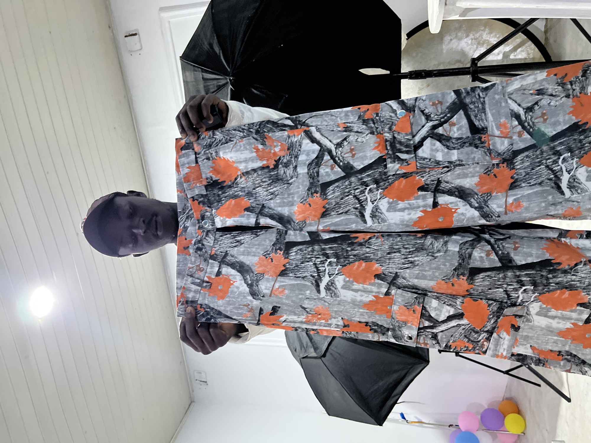 A single male individual, wearing a dark baseball cap and a light-colored long-sleeved shirt, stands frontally in an indoor setting in Unknown, Unknown. He holds up a pair of cargo pants with both hands, presenting them to the viewer. The pants feature a distinctive camouflage pattern composed of gray and black tree bark and branches, accented by scattered vibrant orange maple leaf motifs. Small, metallic grommets or buttons are visible on the waistband and pocket flaps. The background consists of white walls and a white paneled ceiling. A bright circular ceiling light illuminates the scene from above. Two black photography umbrellas or softboxes on stands are positioned on either side of the subject, partially visible. In the lower-left corner, a cluster of deflated colorful balloons (pink, purple, yellow, blue, orange) rests on the floor. A white pedestal or stand is partially visible on the far right. Rectangular fixtures, possibly electrical outlets, are present on the white walls.