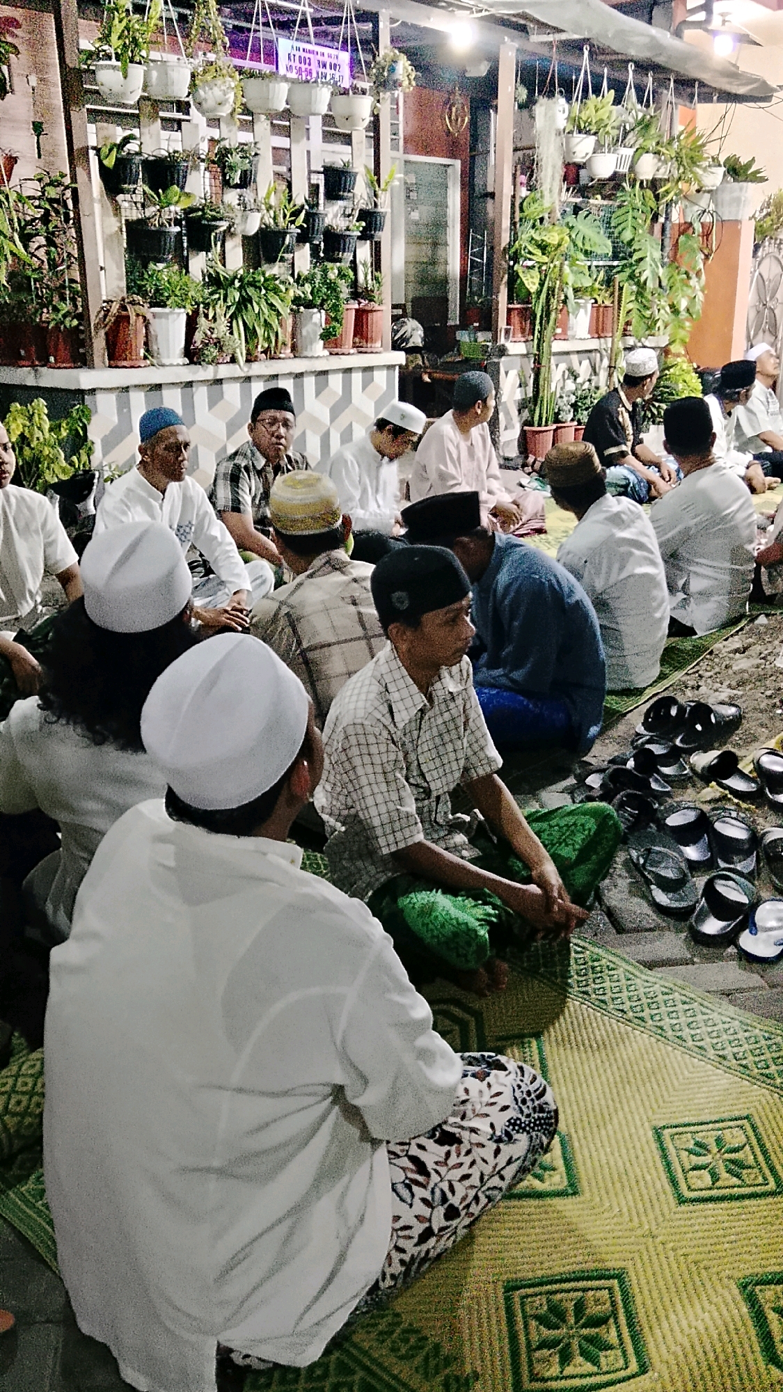 The image depicts a gathering of men, likely engaged in a religious or community event, seated on woven mats on the ground. The setting appears to be outdoors or semi-outdoors, possibly a courtyard or porch area, with potted plants hanging and arranged in the background. The men are dressed in traditional attire, including sarongs, caps (peci), and long-sleeved shirts. Some individuals are wearing white caps, indicative of Islamic faith. The overall atmosphere suggests a communal and possibly solemn occasion. The presence of multiple pairs of footwear suggests that shoes were removed before entering the gathering space. The backdrop includes a building with a patterned wall and a doorway. The lighting is dim, indicating an evening or night-time event. The image is taken from a slightly elevated perspective, looking down upon the seated individuals.