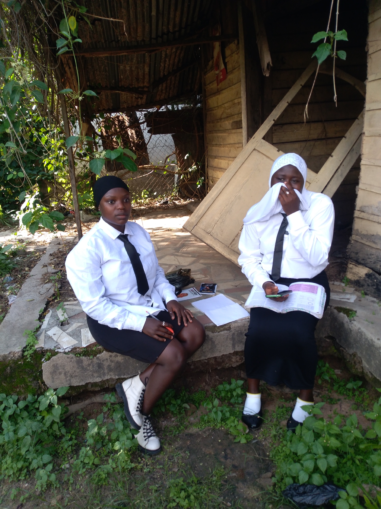 Two young women are seated outdoors on a concrete ledge against a backdrop of dense green vegetation and rustic structures. The specific location within the city of Jos, Nigeria, cannot be determined from the visual information provided.
The woman on the left, with dark skin, wears a black head covering, a white long-sleeved shirt, a black tie, and a black skirt, paired with white and black lace-up boots. She sits with her legs crossed at the ankle, hands resting on her lap, holding a red pen, and looks directly towards the viewer. A ring is visible on her left-hand ring finger.
The woman on the right, also with dark skin, wears a white patterned head covering that partially conceals her face, a white long-sleeved shirt, a black tie, and a black skirt. She wears black shoes with white socks that have a yellow top stripe. She is looking downwards, holding an open book with purple text and a green smartphone in her hands. A ring is visible on her right-hand ring finger.
The immediate surroundings consist of a dirt ground covered with sparse green foliage and weeds. Behind the subjects, on the left, is dense, dark green foliage and what appears to be a corrugated metal roof structure. On the right, a weathered wooden building with light-colored plank siding and exposed framing is visible. Papers and a small black-covered item (possibly a book or CD case) are placed on the concrete ledge between the two women. Another black object, possibly a plastic bag, is on the ground near the right woman's feet. The scene is illuminated by bright sunlight, casting shadows.