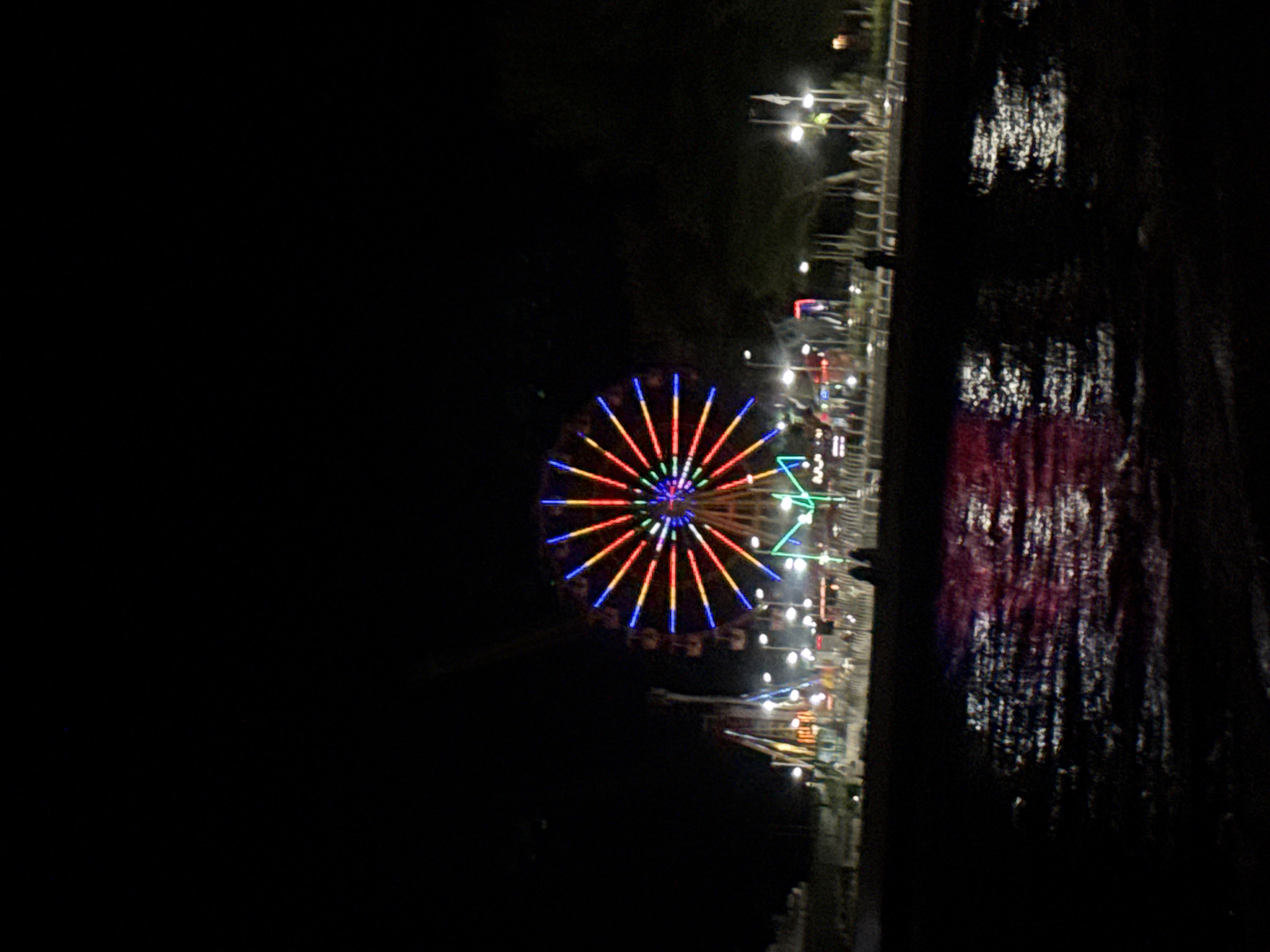 The image depicts a brightly lit Ferris wheel at night in Feodosia, Ukraine. The Ferris wheel is the central and dominant object, adorned with vibrant, multi-colored lights forming radial patterns. To the left of the Ferris wheel, other amusement park rides, also illuminated, are visible. In the foreground, a dark body of water reflects the colorful lights of the Ferris wheel and rides, creating a shimmering, abstract pattern on the surface. Along the shoreline, there are indications of people standing and walking, though they are largely silhouetted against the background lights. The overall scene suggests a lively atmosphere at an amusement park by the water during the evening hours.