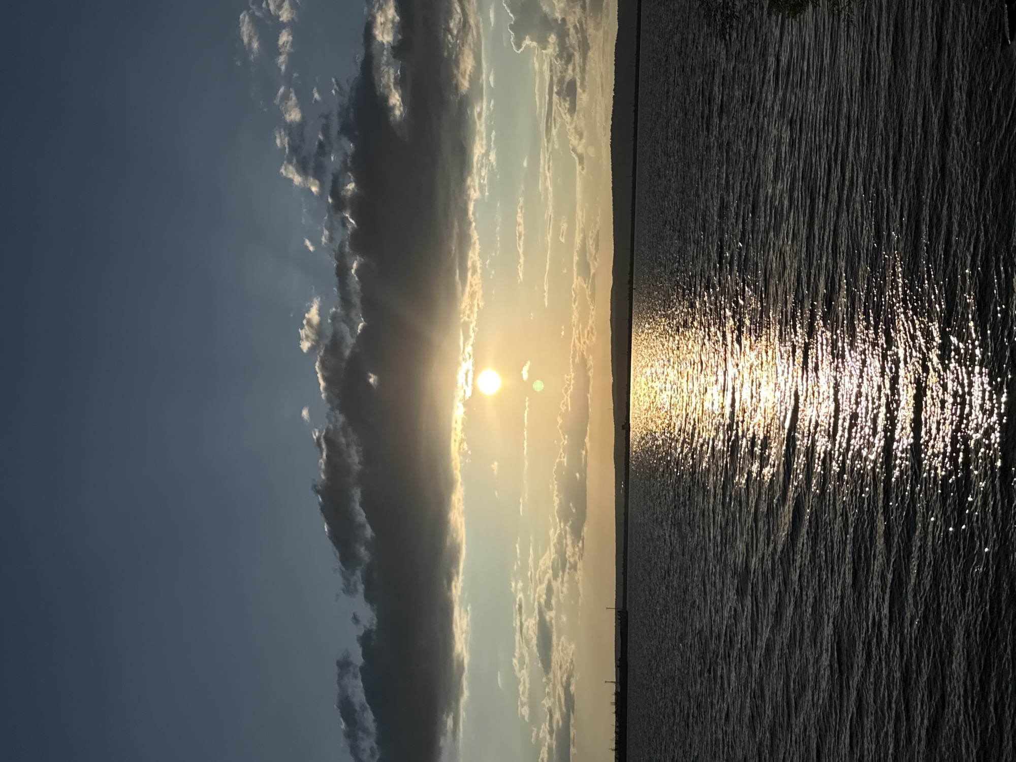 The image depicts a tranquil sunset over the Kama River in Chaikovsky, Russia. The sun, a bright orb, is partially obscured by dark, heavy clouds and rays of light pierce through, casting a golden sheen on the water. The river's surface is rippled, reflecting the sunlight in a shimmering path. In the distance, a faint silhouette of the riverbank with some industrial structures is visible on the left, and a tree line on the right. The sky transitions from a deep blue at the top to lighter shades of yellow and orange near the horizon, with scattered smaller clouds.