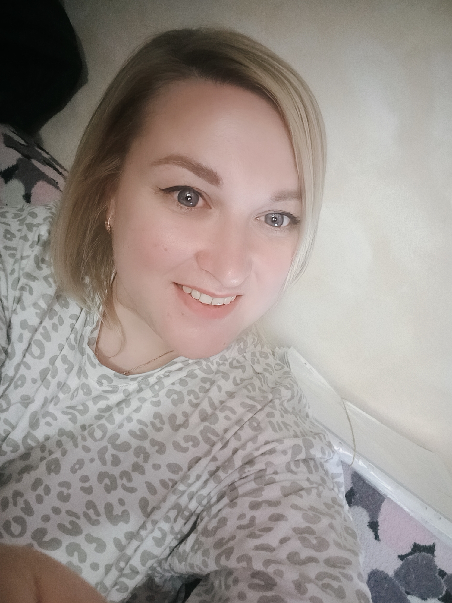 A fair-skinned woman with shoulder-length blonde hair is positioned indoors, smiling and facing the camera directly. She is wearing a long-sleeved shirt with a gray leopard print pattern. A thin gold necklace is visible around her neck, and she is wearing small gold hoop earrings. The background includes a plain, light-colored wall. The lower portion of the frame includes a carpet with a pattern of light pink, gray, and black shapes. Part of what appears to be a bed or other upholstered furniture is visible in the upper left corner. Location: Unknown, Unknown.