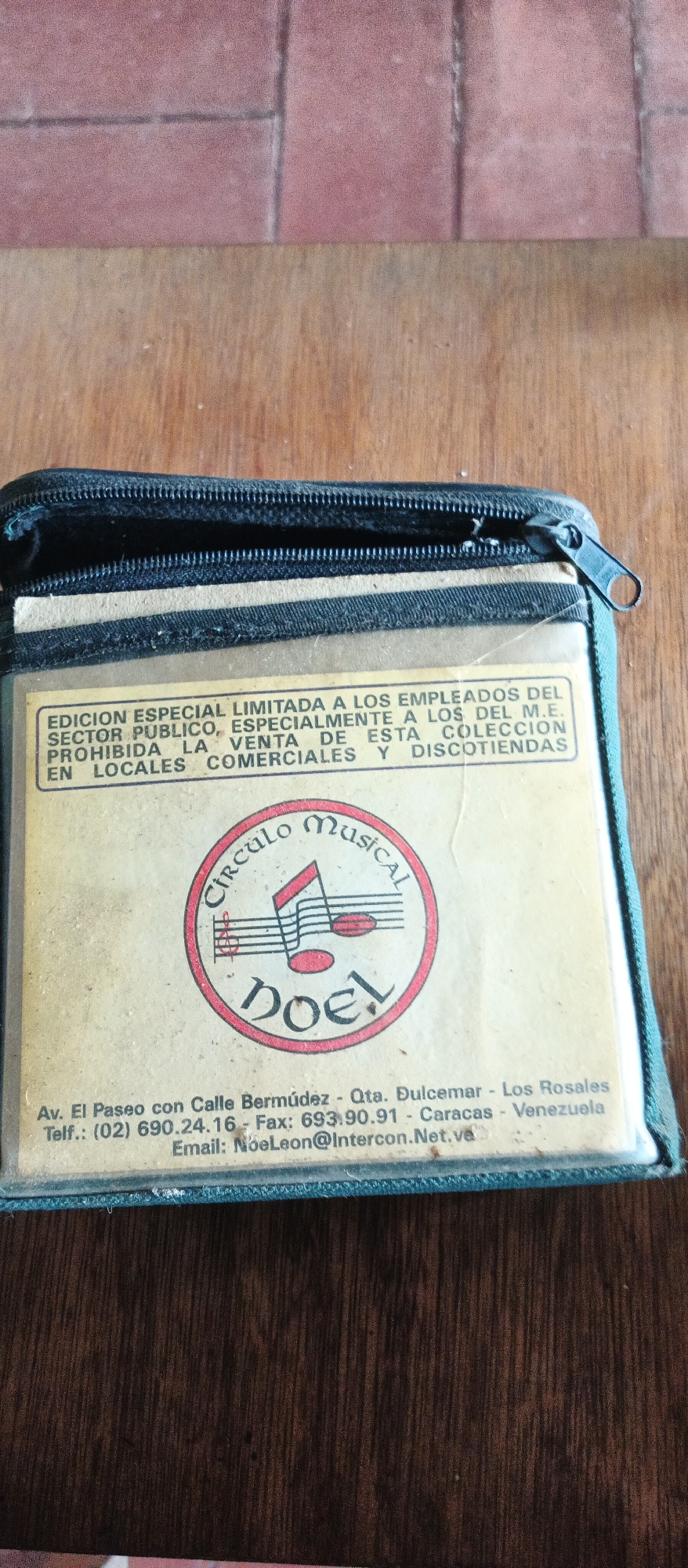 This is a close-up, top-down view of a wallet or case holding what appears to be a CD or cassette tape. The case is dark green and partially unzipped, revealing a light-colored insert with text and a circular logo. The insert has a yellow background and features a red and black circular logo with musical notes and the words "Circulo Musical" and "DOEL" in stylized font. Below the logo, there is text with contact information, including an address in Caracas, Venezuela, a phone number, a fax number, and an email address. The text on the insert also states that this is a special limited edition for employees of the public sector and that its sale in commercial establishments and record stores is prohibited. The background of the image is a wooden surface.