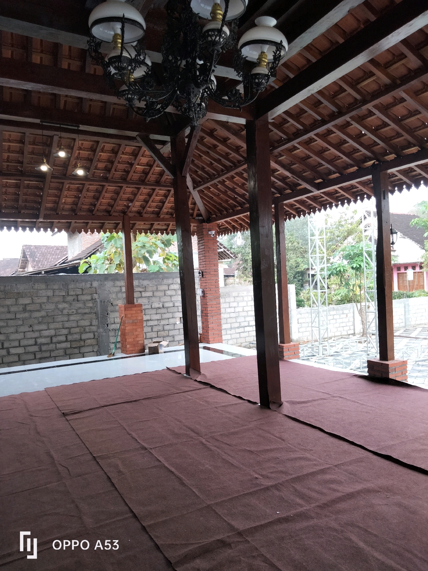 The image captures a covered outdoor space with a traditional Indonesian architectural style, possibly a veranda or an open-air pavilion, located in the city of Trenggalek, Indonesia. The structure features a dark wooden beam ceiling with terracotta tiles, supported by dark wooden pillars that rest on brick bases. Hanging from the ceiling are two ornate chandeliers with white glass shades and decorative dark metalwork. The floor is covered with large pieces of dark brown fabric, appearing to be carpeting or a protective covering, laid out unevenly.
In the background, a grey brick wall forms a boundary, with some lighter colored plastering visible. Beyond the wall, trees and houses with terracotta roofs suggest a residential or semi-rural setting. A metal grate structure is also visible in the distance, along with a security camera mounted on a brick pillar. The scene is well-lit, likely due to natural daylight, casting subtle shadows. There are no people present in the image. The overall impression is one of a serene, traditional outdoor area, possibly awaiting an event or in the process of being set up.