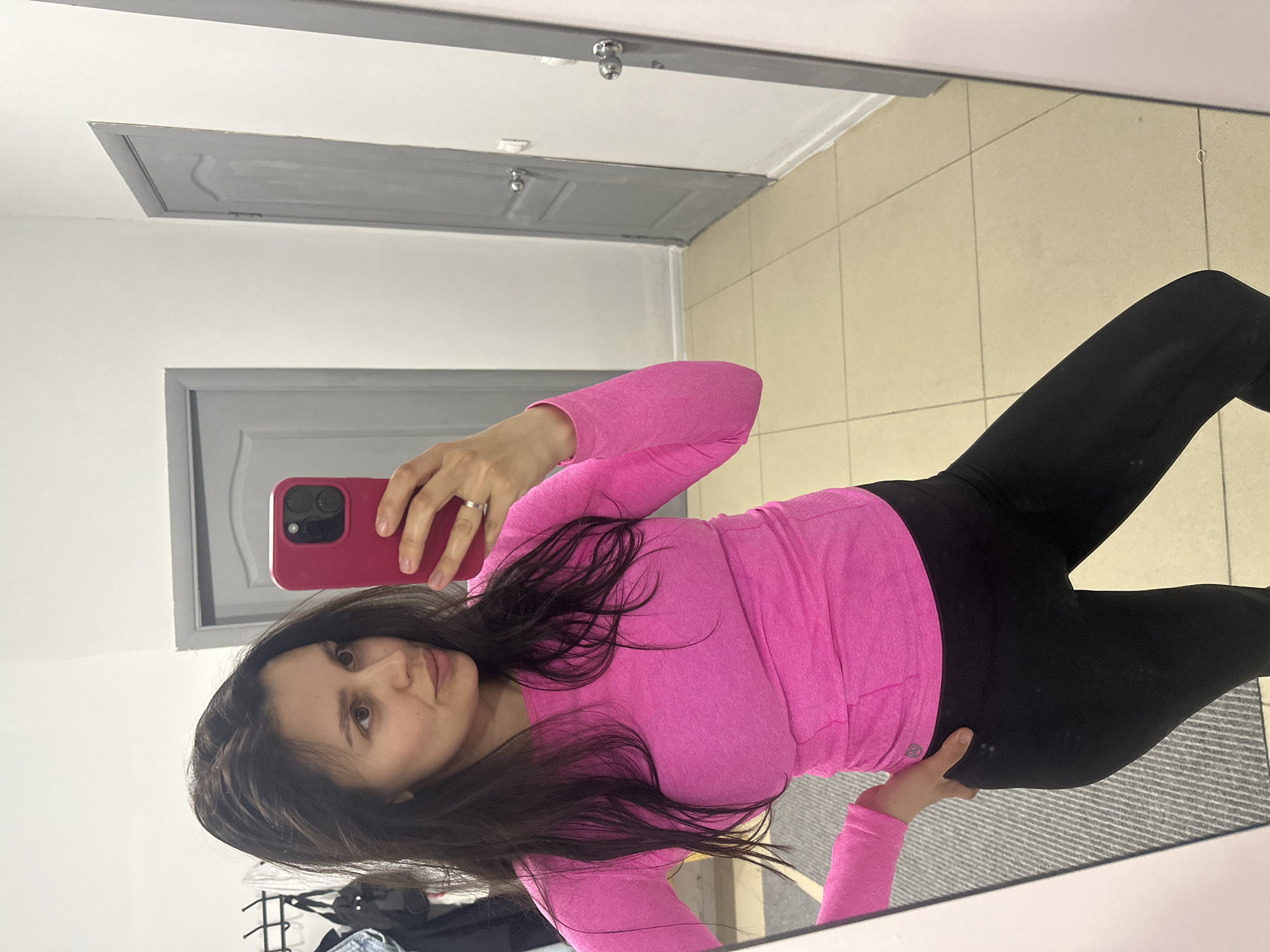 A woman takes a mirror selfie indoors, appearing rotated 90 degrees clockwise in the original image. She has long, dark hair and is wearing a form-fitting, long-sleeved hot pink top and black leggings. A small, white circular logo (resembling a brand logo like Lululemon) is visible on her left hip area. She holds a smartphone with a red/pink case in her right hand, positioned to capture her reflection, and has a ring on her left ring finger. Her left hand is resting on her hip.
The background, visible as a reflection in the mirror, features plain light-colored walls and several gray, paneled interior doors. One door or access panel is partially visible above her head. The floor is covered with light-colored square tiles, and a gray striped rug is seen near her feet. The overall setting suggests an indoor space, possibly a changing room or a well-lit hallway. No specific time of day or weather can be determined. The woman has a neutral expression as she looks towards the camera. No discernible text is clearly visible other than the logo on her top. The location context provided is Chaikovsky, Russia.