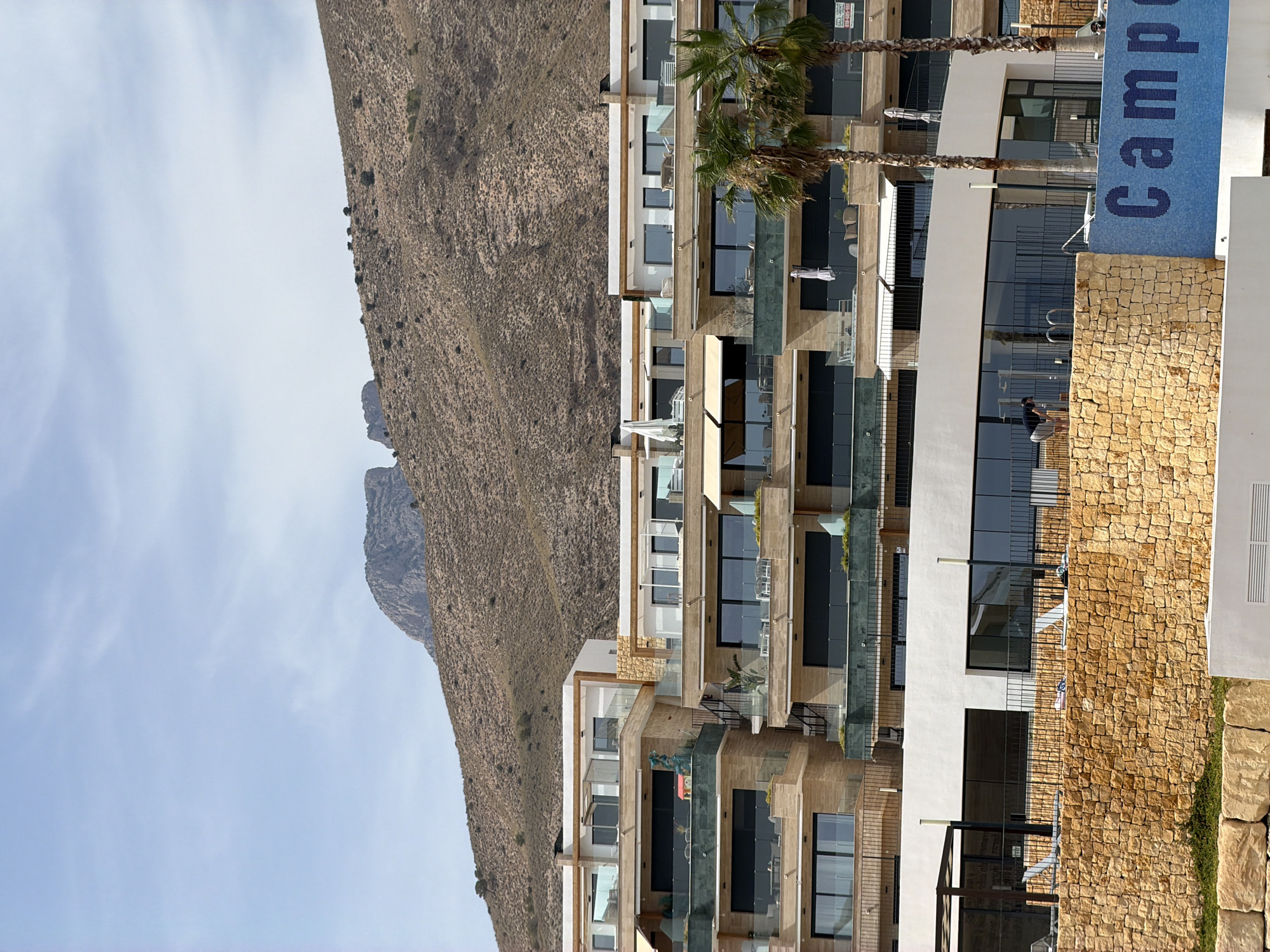 In Finestrat, Spain, a multi-story modern building complex is captured against a backdrop of a rugged, arid mountain range. The building features multiple levels with balconies and large windows, many with dark tinted glass. The architecture appears contemporary, with a combination of light-colored stone, white facades, and glass railings. Two palm trees stand in the foreground to the right, their fronds partially obscuring the building.
Below the main building structure, a lower section of the complex has a white facade and large glass windows, suggesting a communal area or reception. A section of this lower building is adorned with blue mosaic tiles that spell out "Camp". A low, light brown stone wall separates the lower building from an area with white lounge chairs, implying a pool or relaxation zone. A person in dark clothing and light shorts is seen bending down near this wall, facing away from the camera.
The mountain behind the building is dry and rocky, with sparse vegetation. The sky is a pale blue with wispy white clouds, indicating a clear, possibly warm day. The overall scene depicts a resort or residential development situated in a Mediterranean coastal landscape, characterized by its proximity to natural terrain.