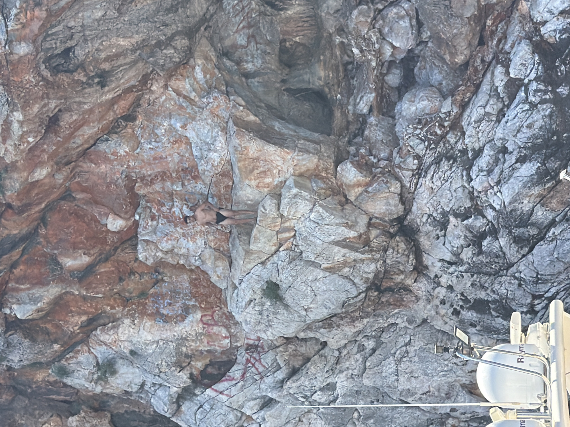 This image captures a rugged, rocky cliff face along the coastline, likely near Alanya, Turkey, under bright daylight. The original image appears rotated, showing the cliff as a prominent vertical feature.
A single person, seemingly a man wearing dark swim trunks, is visible lying flat on a relatively horizontal ledge of the rock face. He appears to be resting or sunbathing. The cliff itself is composed of stratified rock, showcasing a variety of textures and colors including shades of grey, brown, and reddish-brown, with some lighter patches.
Several areas on the rock show signs of human interaction beyond the person, specifically graffiti or painted markings in red and possibly faded blue.
In the lower right portion of the frame, parts of a white boat or yacht are visible, indicating that the photo was taken from the water. Visible boat equipment includes a white spherical dome, likely a radar or satellite antenna, with the text "Rayma" (suggesting "Raymarine") partially legible on its surface, along with metal railings and other nautical instruments. The scene suggests a leisurely boat trip exploring a scenic, rocky coast on a clear, sunny day.