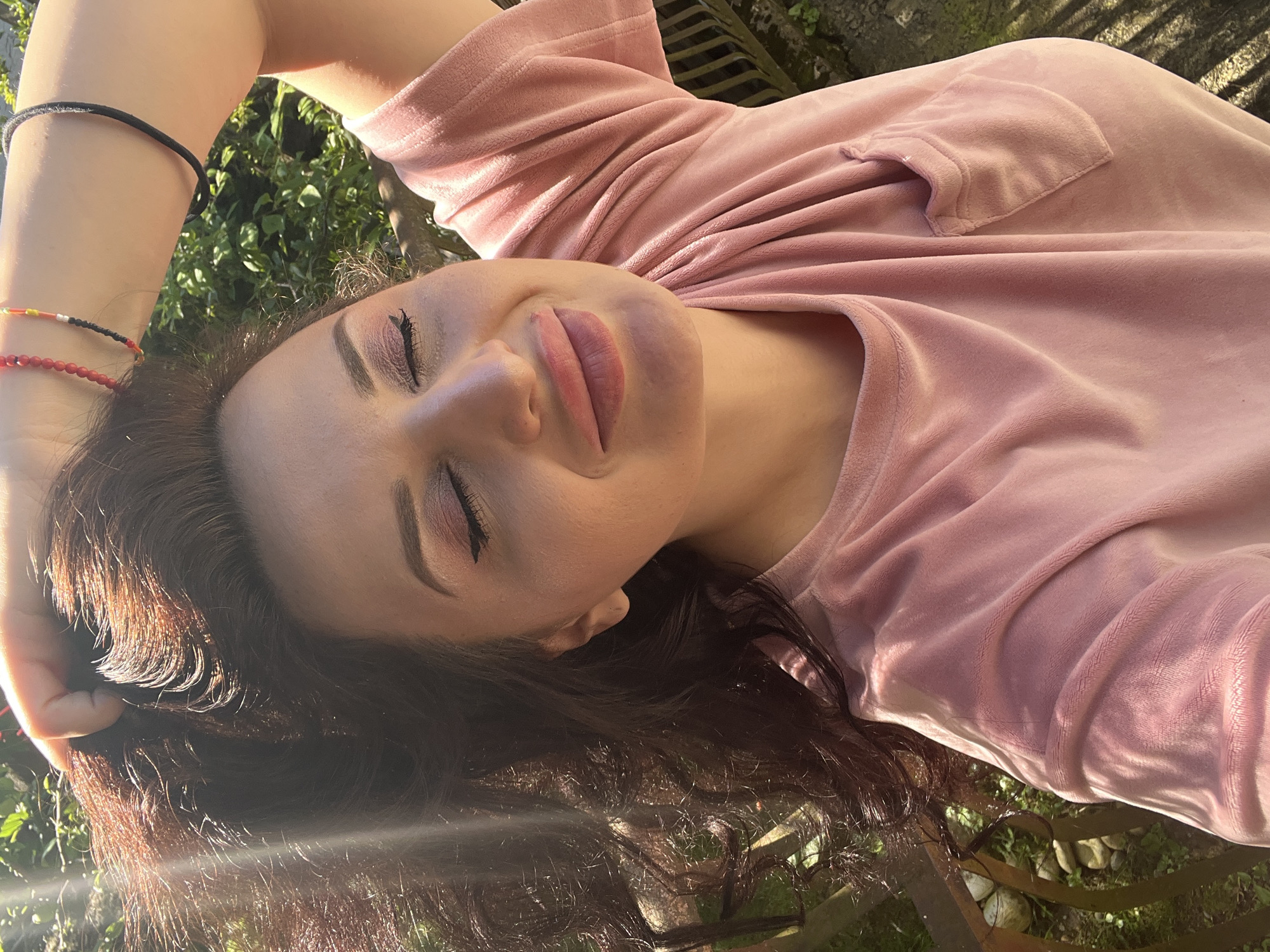 An overhead shot, taken at an angle, captures a person (likely a woman) lying down outdoors, seemingly relaxed under natural light. Her head is tilted, and her eyes are closed, with her lips slightly parted. She has dark hair, which is spread out around her head, and is wearing a soft, velvety pink top with a small pocket detail on the upper right side. Makeup is visible on her face, including pink/purple eyeshadow and eyeliner, and well-defined eyebrows.
On her left wrist, she wears several bracelets: one black cord, one made of red beads, and another multi-colored beaded bracelet featuring red, orange, and black elements. The background features lush green foliage, possibly grass and plants, suggesting a natural outdoor setting. There are also visible elements that appear to be part of a wooden fence or bench, and some small rocks on the ground. Sunlight streams into the scene, creating bright highlights on her hair and skin, and producing lens flare effects, indicating a sunny day, possibly morning or late afternoon. The overall impression is one of peacefulness and calm. No text is visible in the image.
